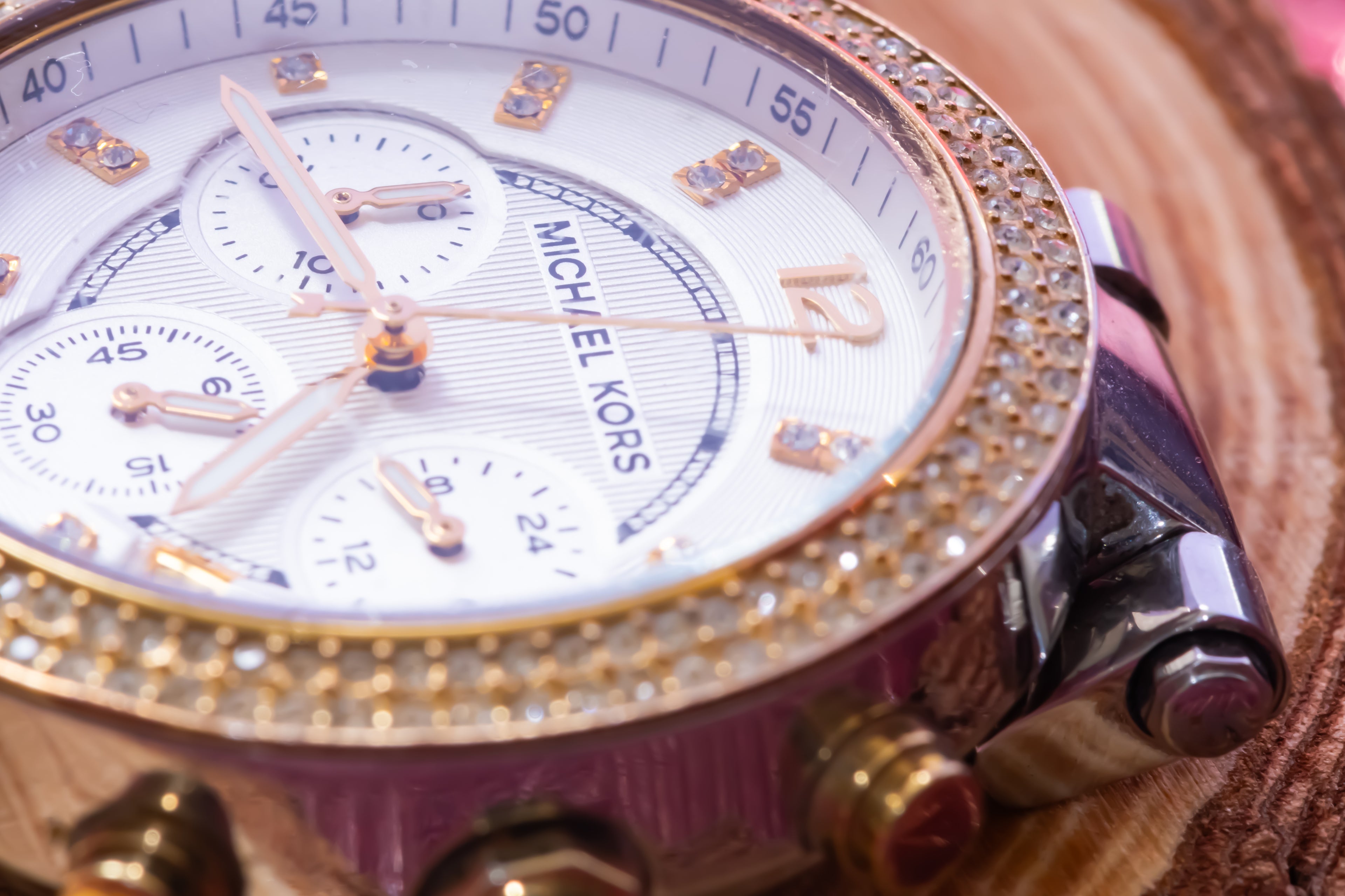 A close-up of a Michael Kors watch featuring a white dial, gold-tone bezel, and pink-toned crystal embellishments, resting on a textured brown surface.