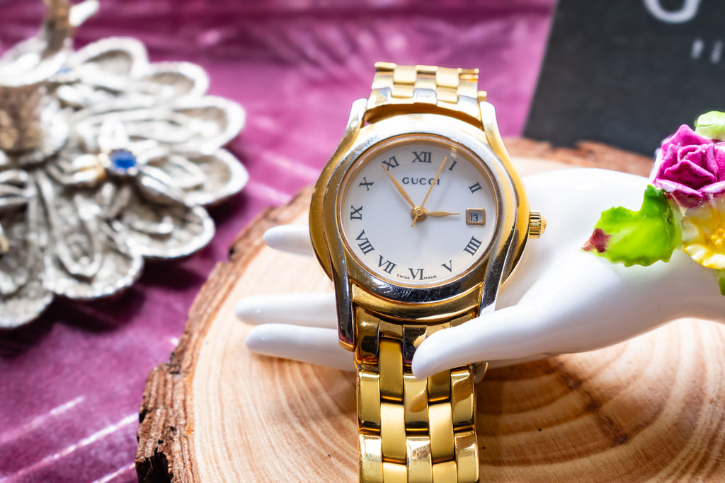 A Gucci gold-tone metal watch with a white dial and Roman numeral hour markers, displayed on a white hand-shaped stand over a wood slice with decorative flowers and a purple fabric background.