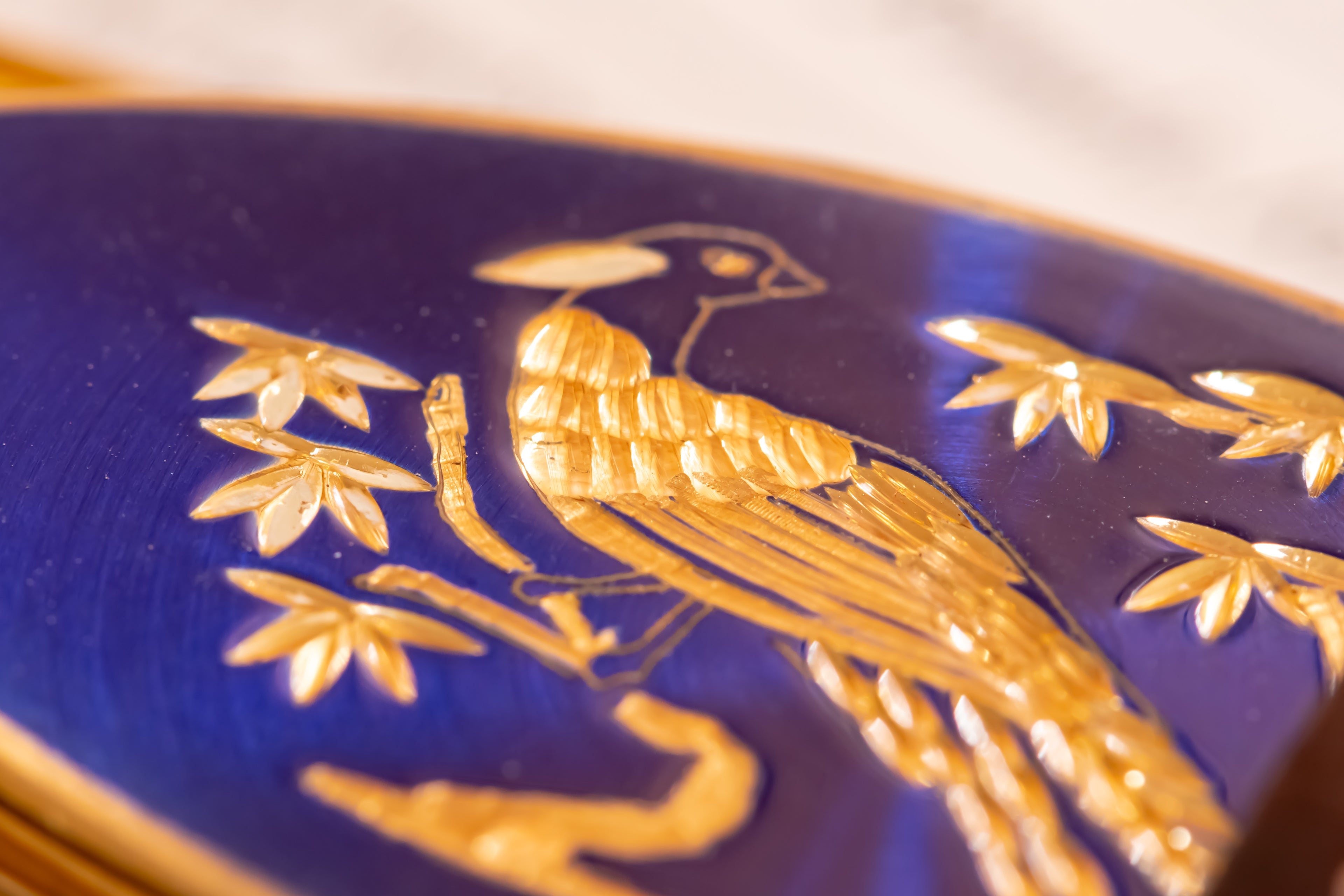 Close-up of a dark blue oval lipstick case featuring raised gold relief decoration of a bird perched among leaves.