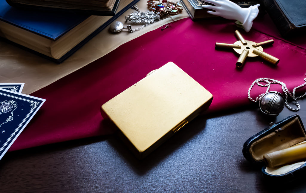 A square gold-toned metal case or compact lies on a dark wooden table, surrounded by occult-themed props including a deep red cloth, old books, crystals, a small white skull, a brass key, and an open pocket watch.