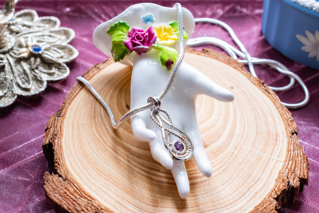 A silver teardrop pendant necklace with a purple gemstone, displayed on a white ceramic hand-shaped stand decorated with small flowers, set on a wood slice over a purple fabric background.