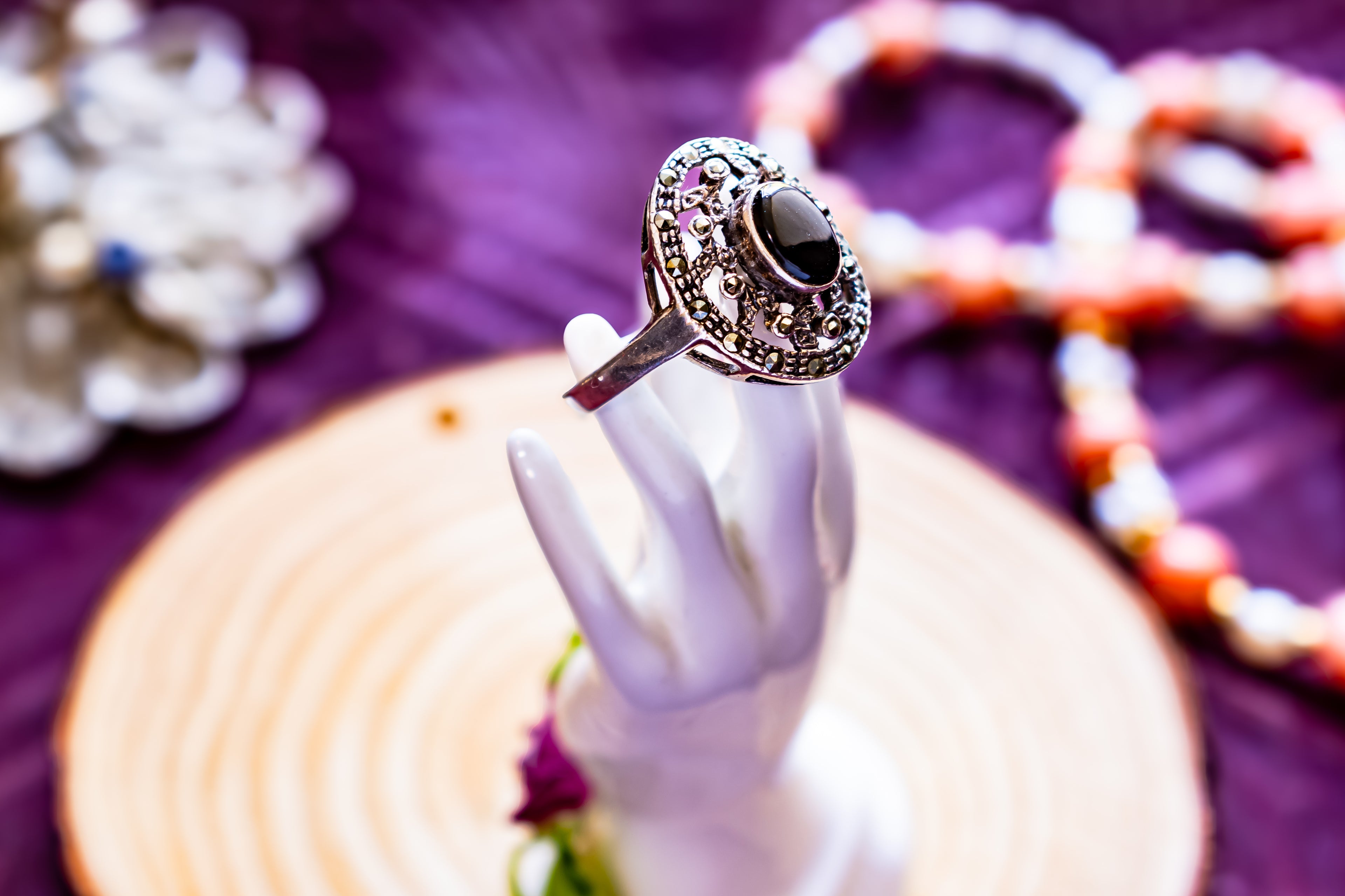 A silver-toned statement ring with a large black oval center stone and surrounding pink and clear stones, displayed on a white hand-shaped stand over a wooden slice on a purple fabric background.