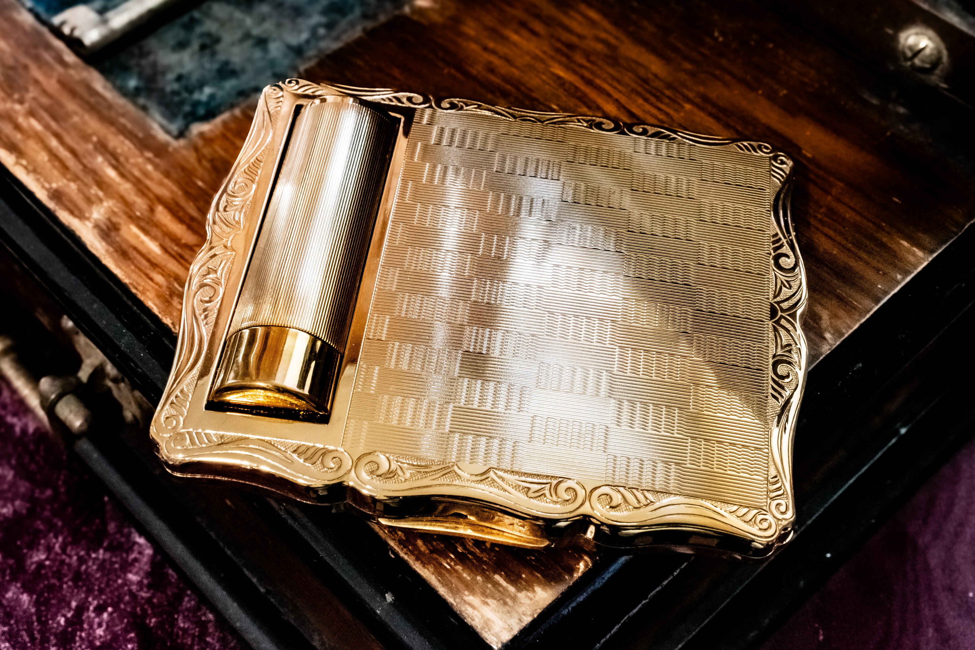 An ornate gold-toned metal square powder compact with a textured patterned center and decorative border, shown resting on a dark wooden surface.