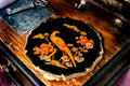 An ornate black compact mirror featuring a gold bird and floral motif, displayed on a vintage wooden box with a key nearby.