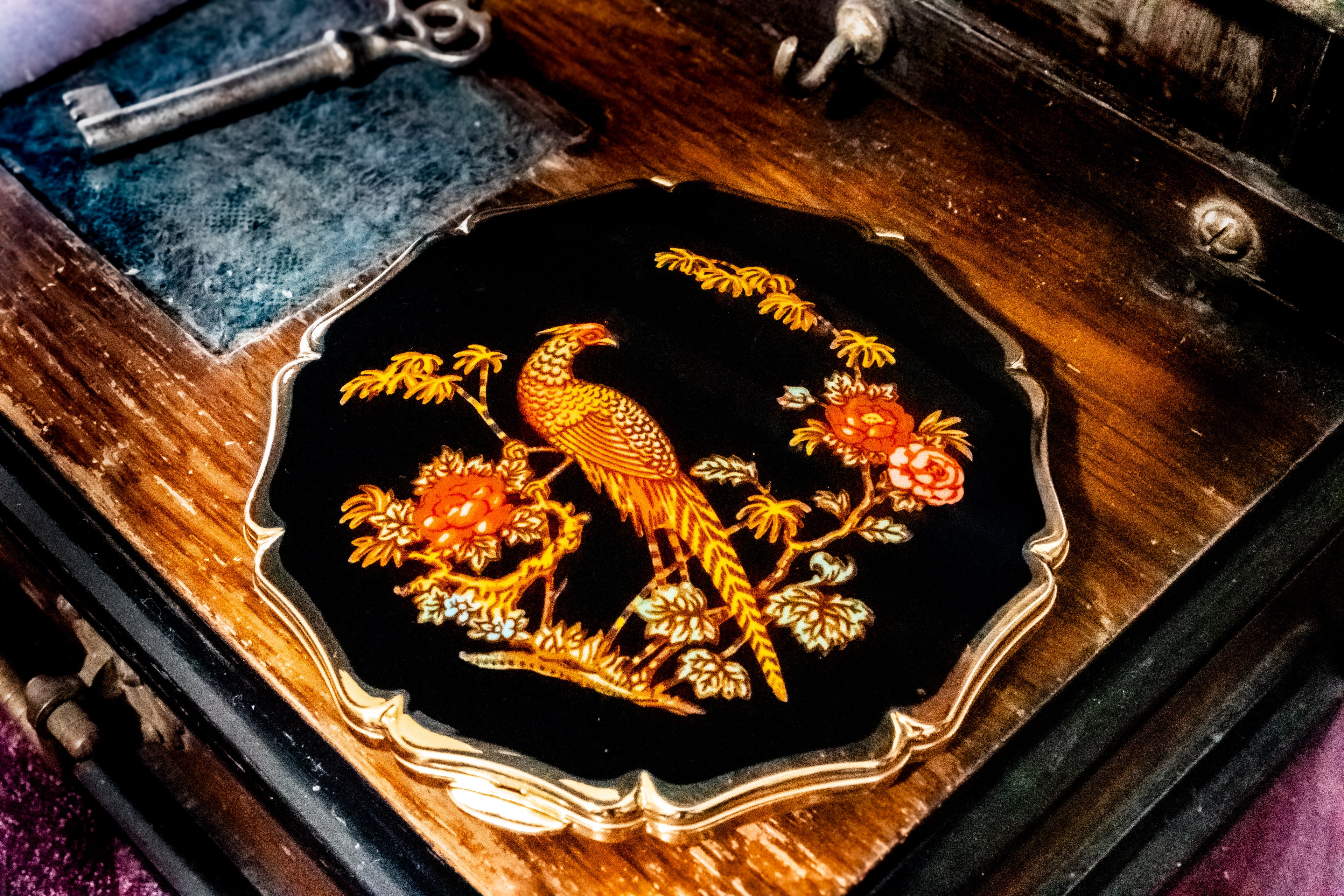An ornate black compact mirror featuring a gold bird and floral motif, displayed on a vintage wooden box with a key nearby.