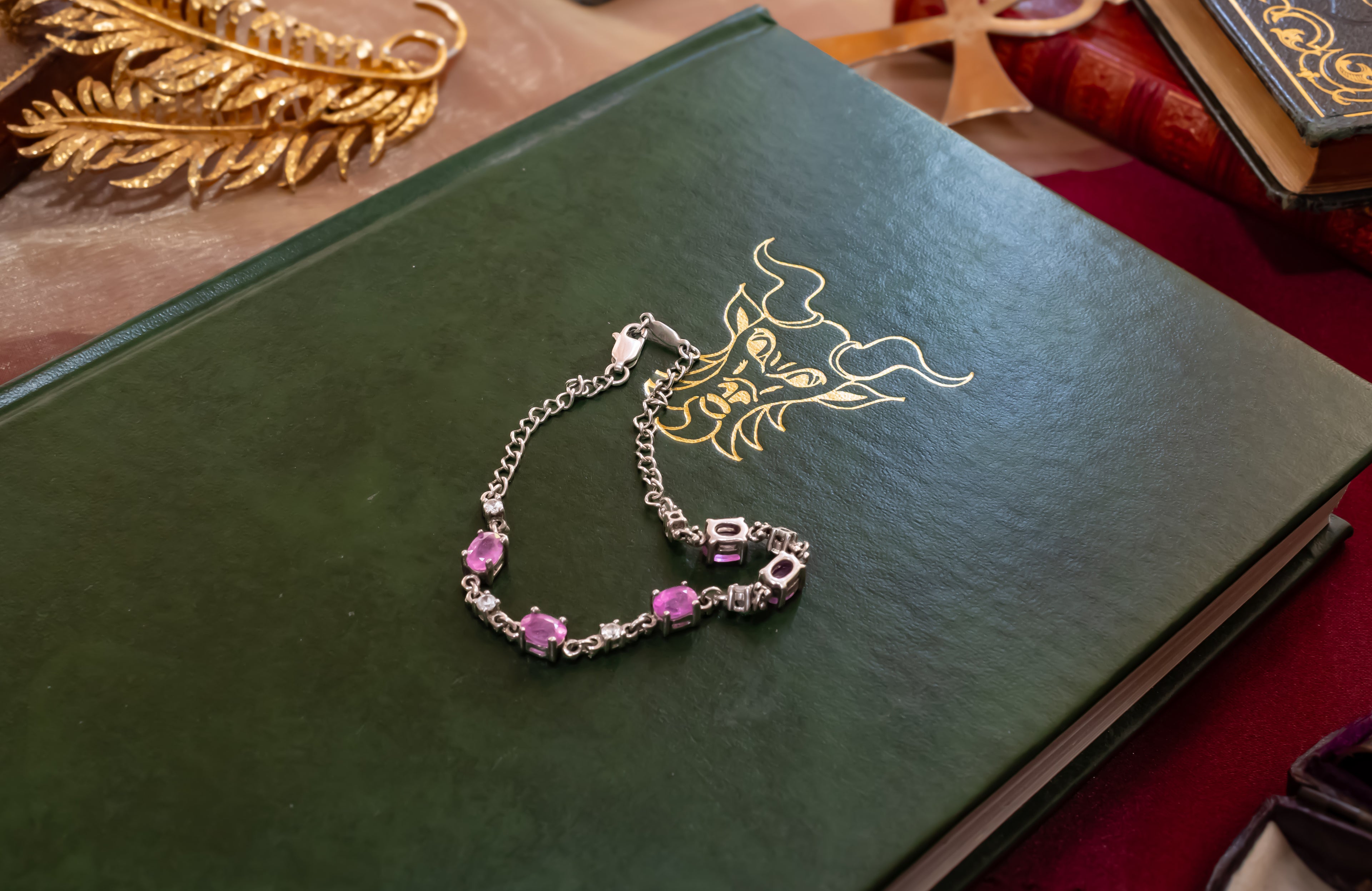 A silver chain bracelet with pink gemstones and a heart-shaped charm, displayed on a dark green book with gold detailing.