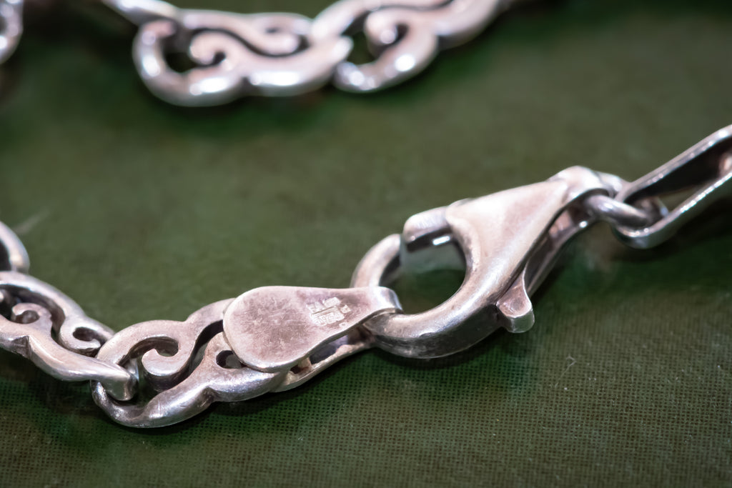 Close-up of a silver-toned chain bracelet with decorative scrollwork links and a lobster clasp, resting on a dark green textured surface.