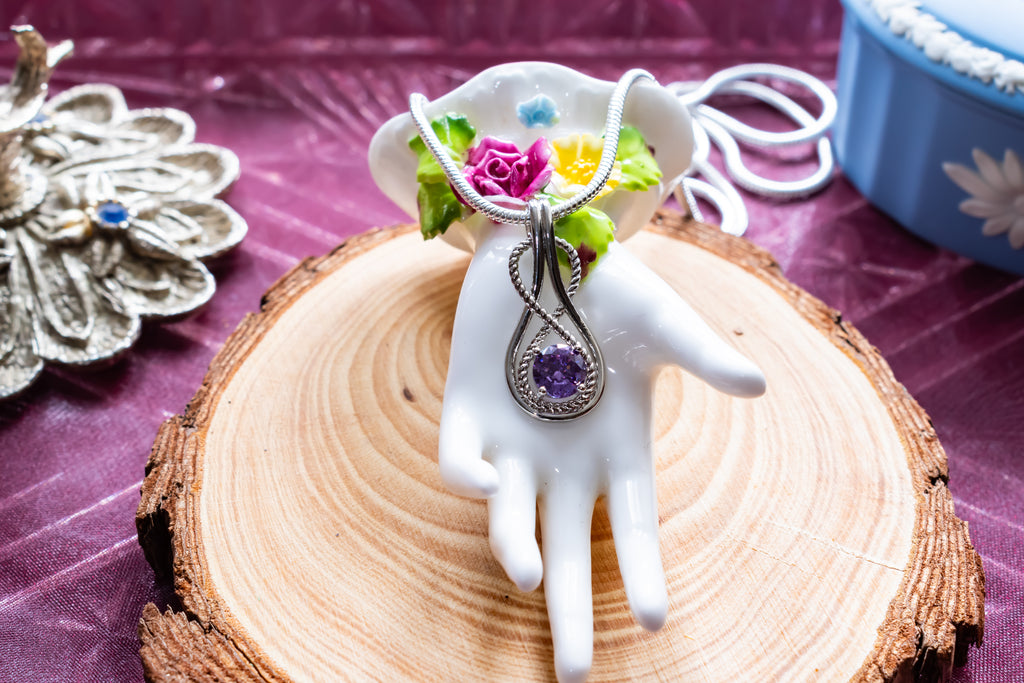 A silver pendant necklace with a purple teardrop gemstone is displayed on a white hand-shaped mannequin. The necklace is decorated with small artificial flowers and green leaves, resting on a rustic wood slice over a purple fabric background.