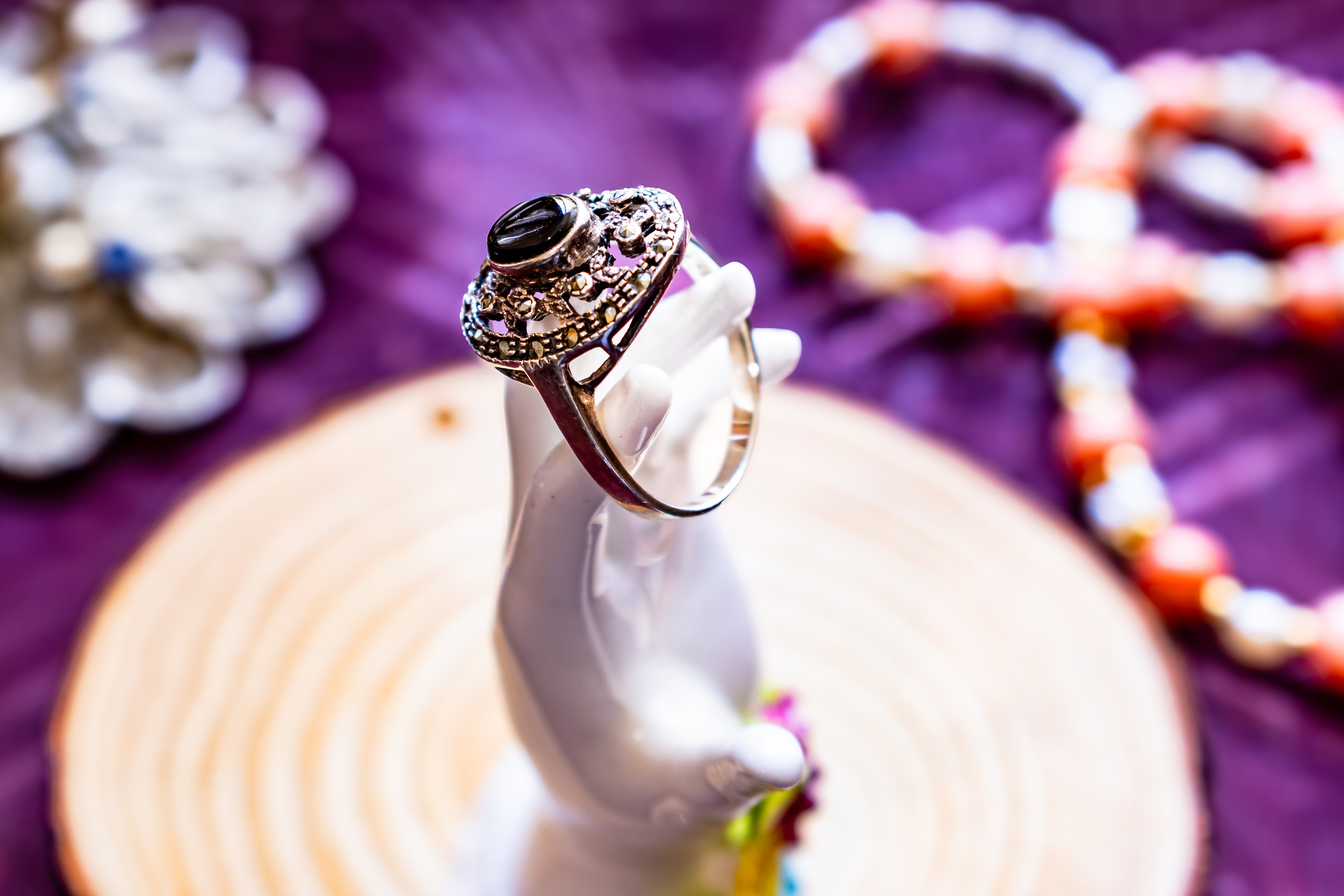 An ornate silver ring with a black oval stone and pink gemstone accents, displayed on a white hand-shaped stand. The background features a purple fabric backdrop with blurred coral and white beaded jewelry.