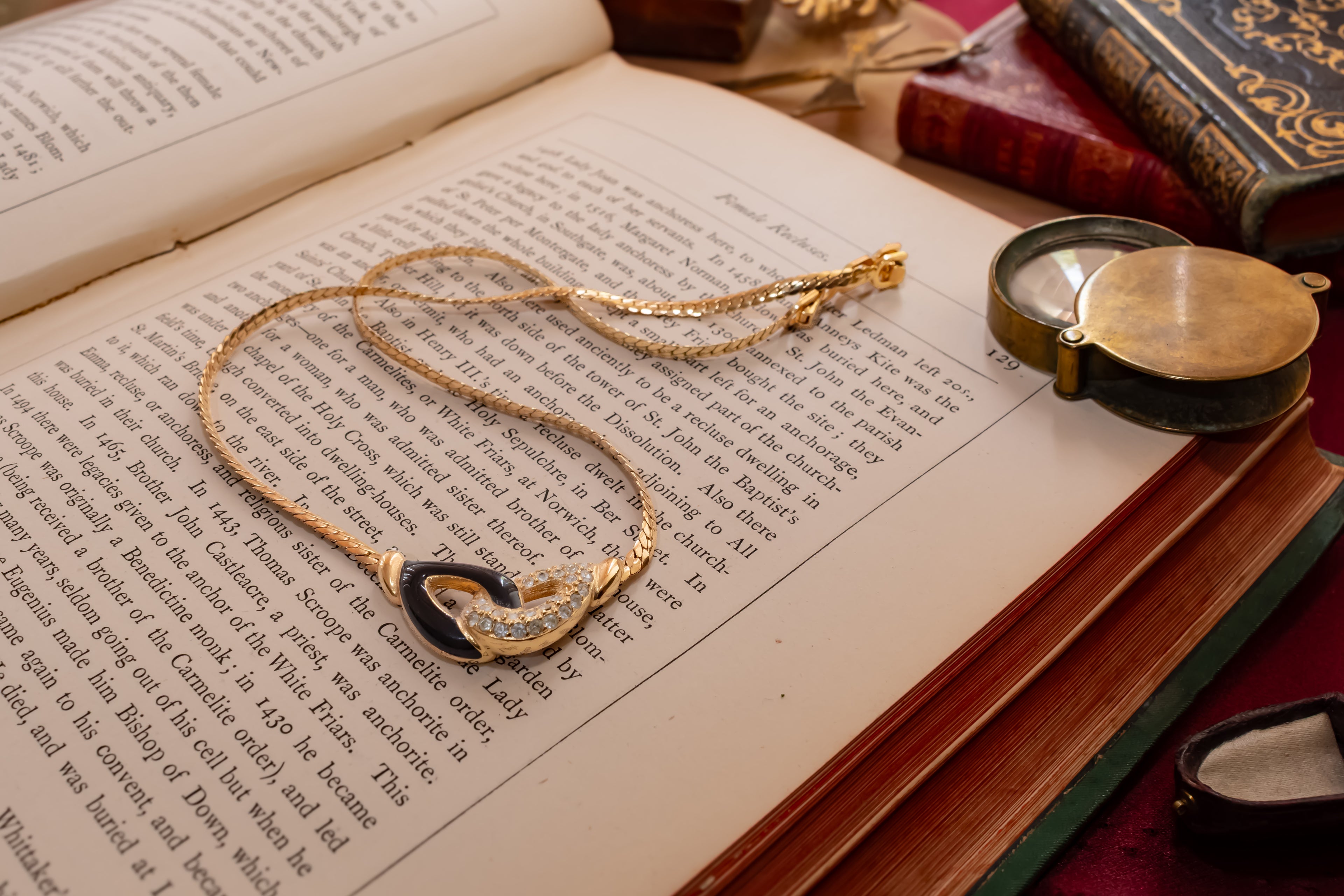 A Christian Dior gold chain necklace with a black heart-shaped pendant and small clear stones, displayed on an open vintage book next to a brass pocket watch.