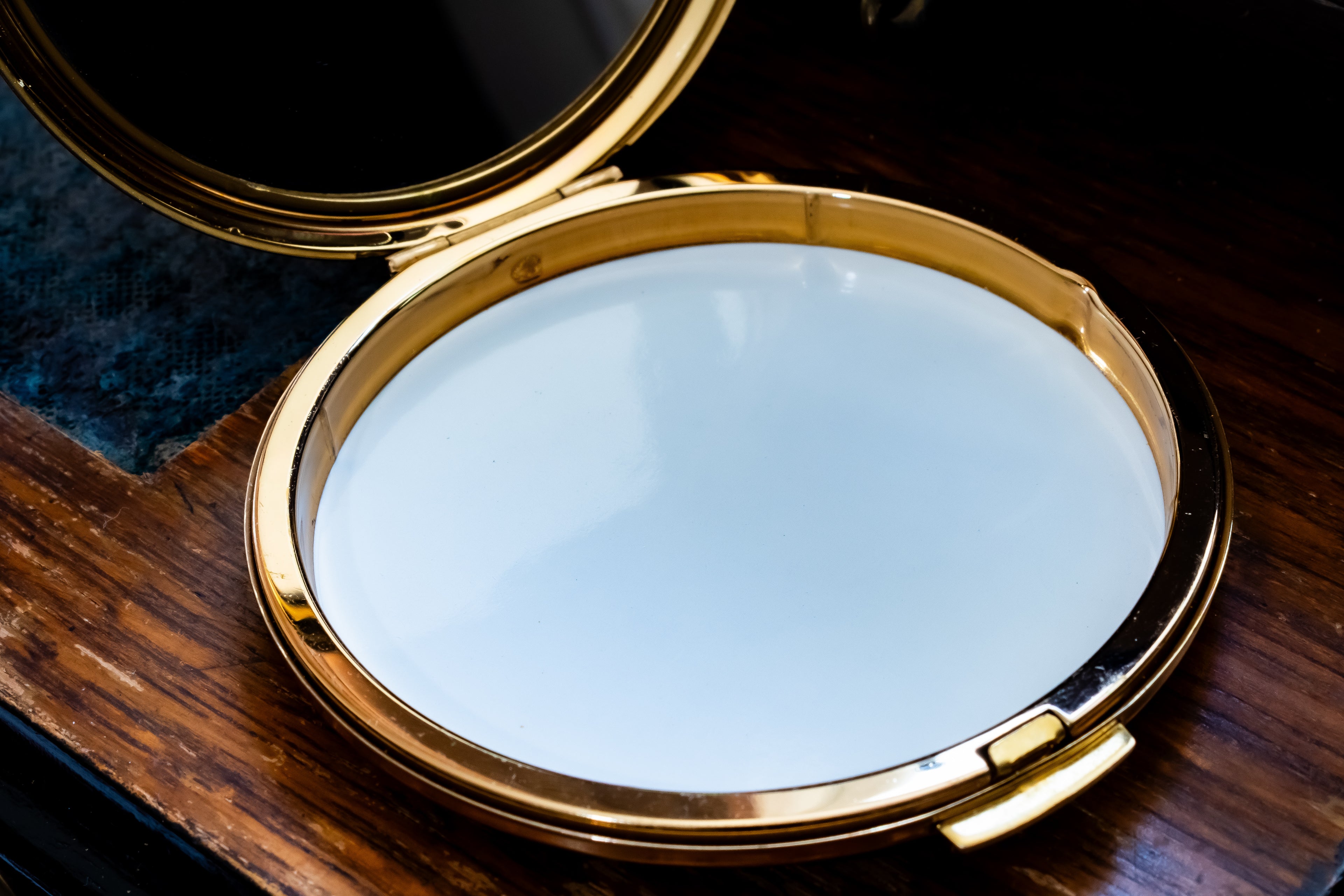 An open round compact mirror with a polished gold-tone frame resting on a dark wooden surface.