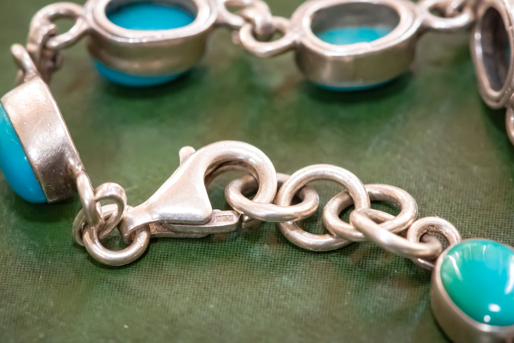 A close-up of a silver chain bracelet with round turquoise stones, showing a lobster clasp and the links against a textured green background.