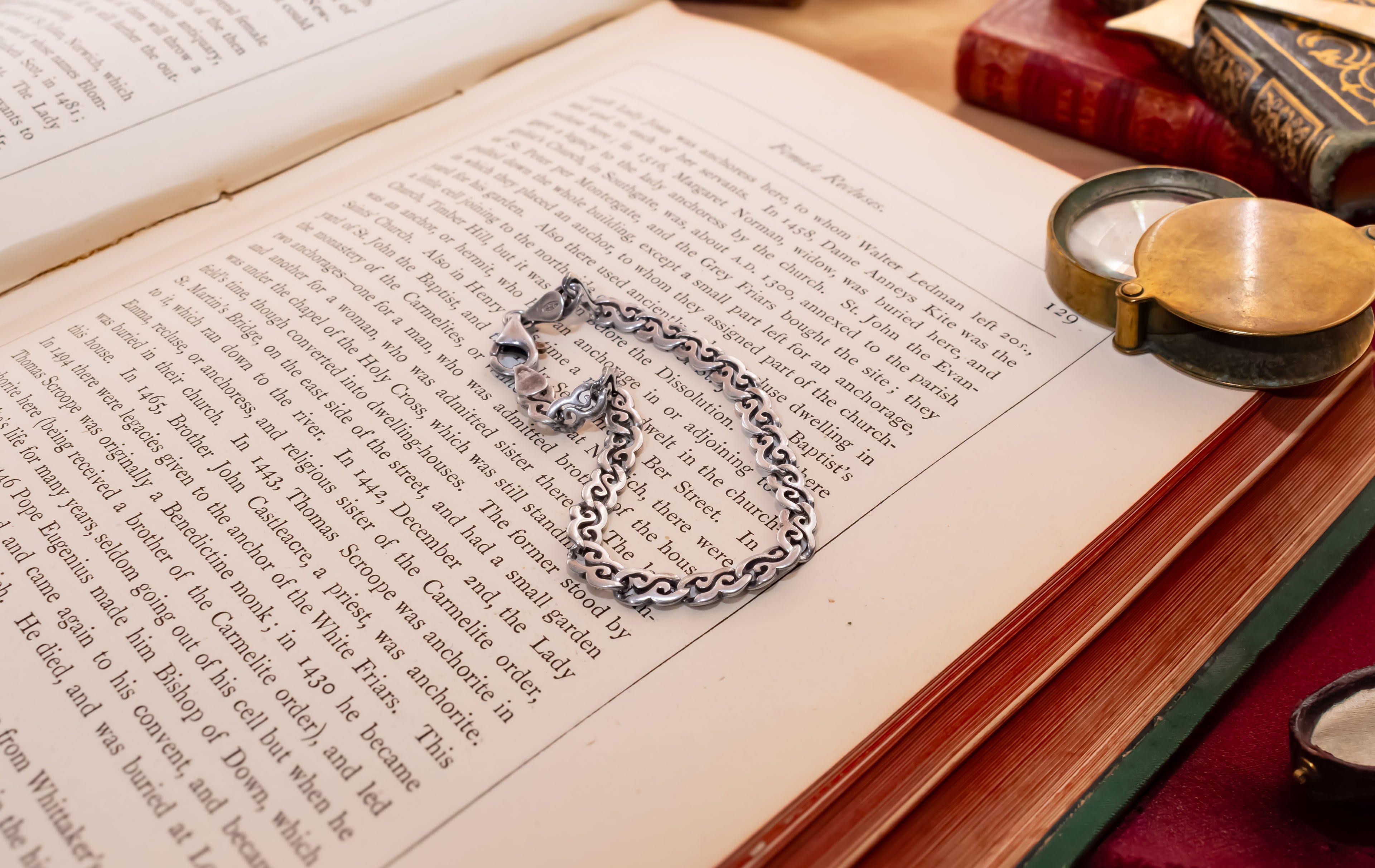 A silver-toned chain necklace with a small heart-shaped pendant rests on the pages of an open vintage book, surrounded by old books and a brass pocket mirror on a wooden surface.
