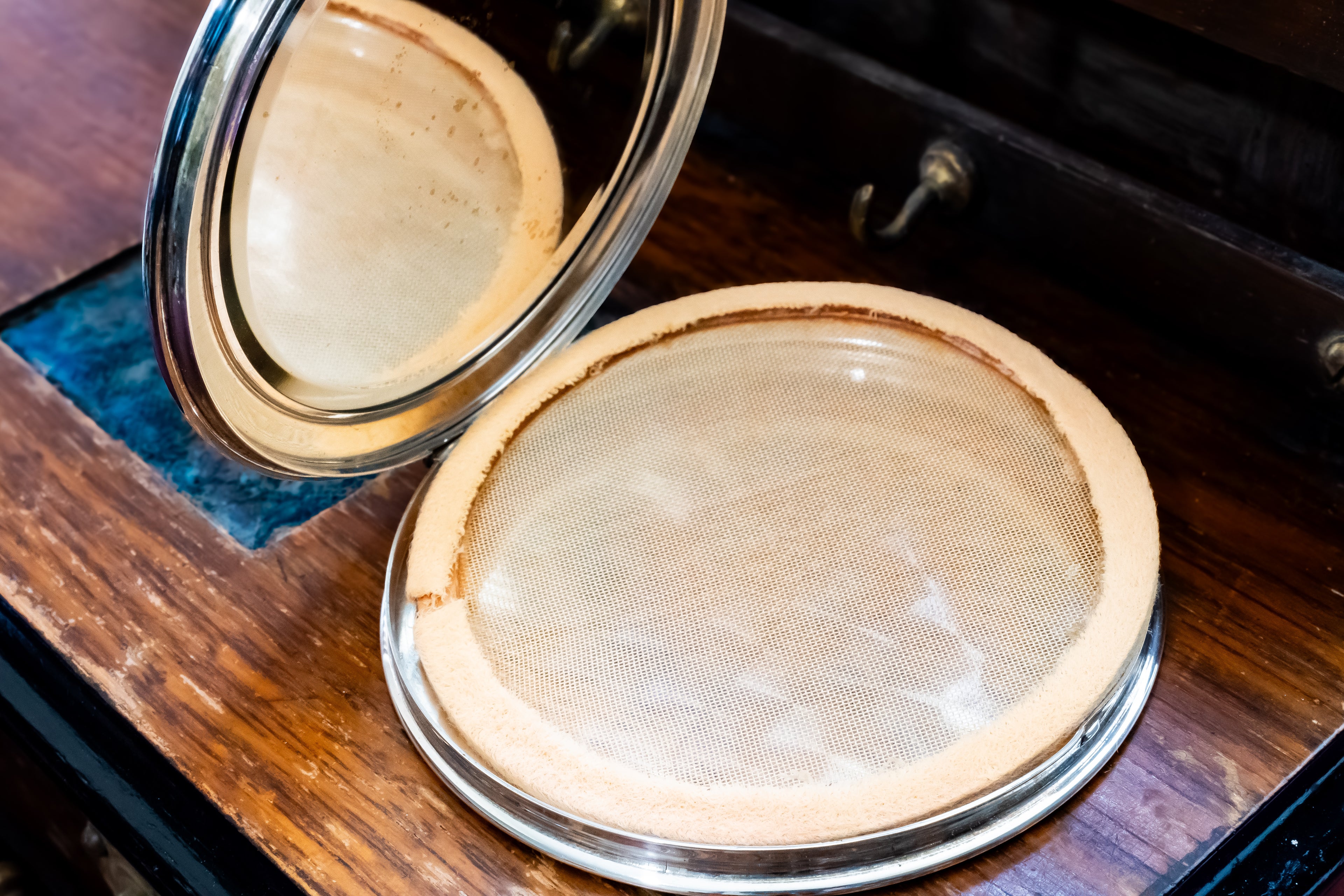 An open round vintage compact mirror with a silver metal frame and a beige mesh screen, resting on a dark wooden surface.
