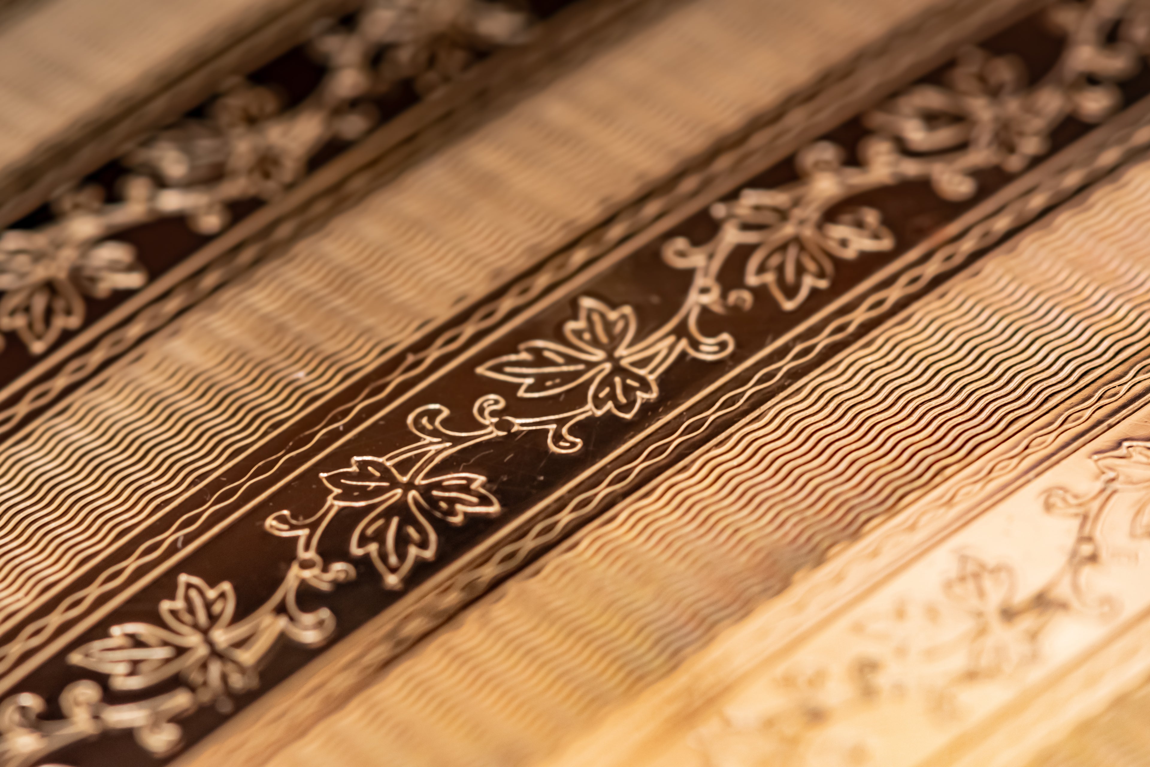 Close-up of a powder compact with repeating horizontal bands in warm beige, tan, and dark brown, featuring floral and vine motifs.