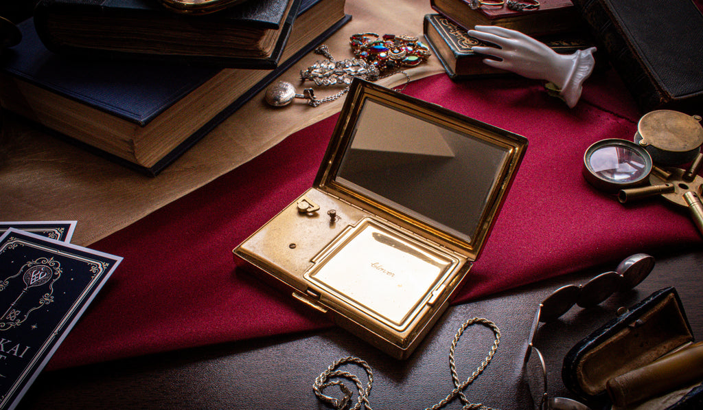An open gold-toned vintage compact or case with a mirror and small inset compartment, displayed on a red cloth surrounded by old books, jewelry, cards, and other antique props.