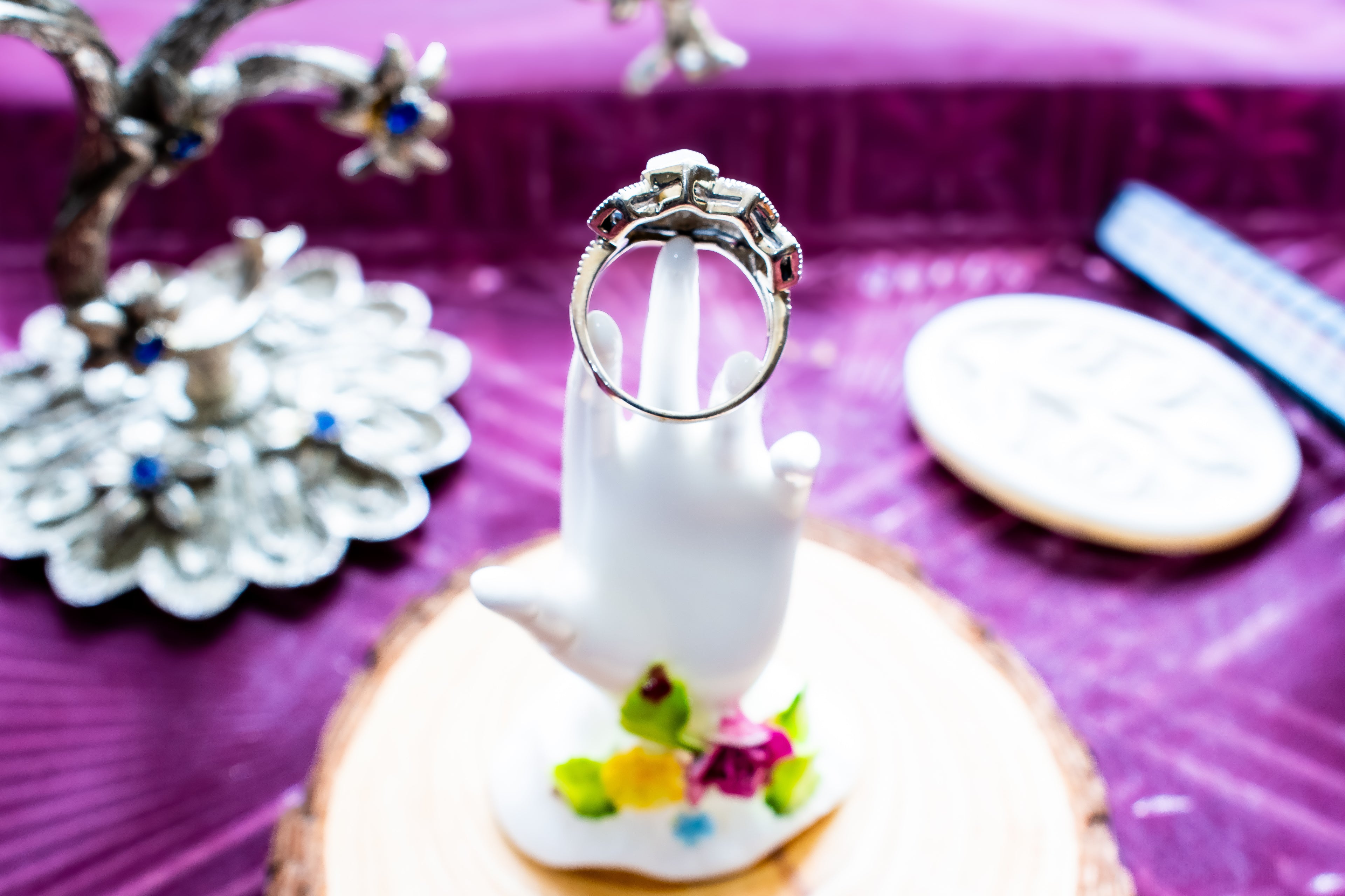 A silver ring displayed on a white hand-shaped jewelry stand, set on a round wood slice with a purple fabric background and blurred decorative jewelry nearby.