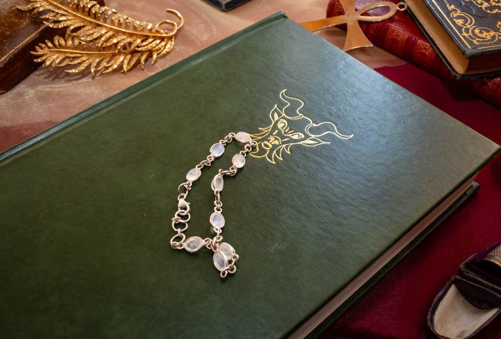 A silver-toned chain bracelet with round white stones and a small dangling charm, laid on top of a dark green book with gold lettering.