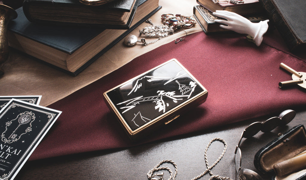 A rectangular black and gold case with an illustrated cover design, styled on a dark table alongside books, cards, jewelry, and other vintage props.