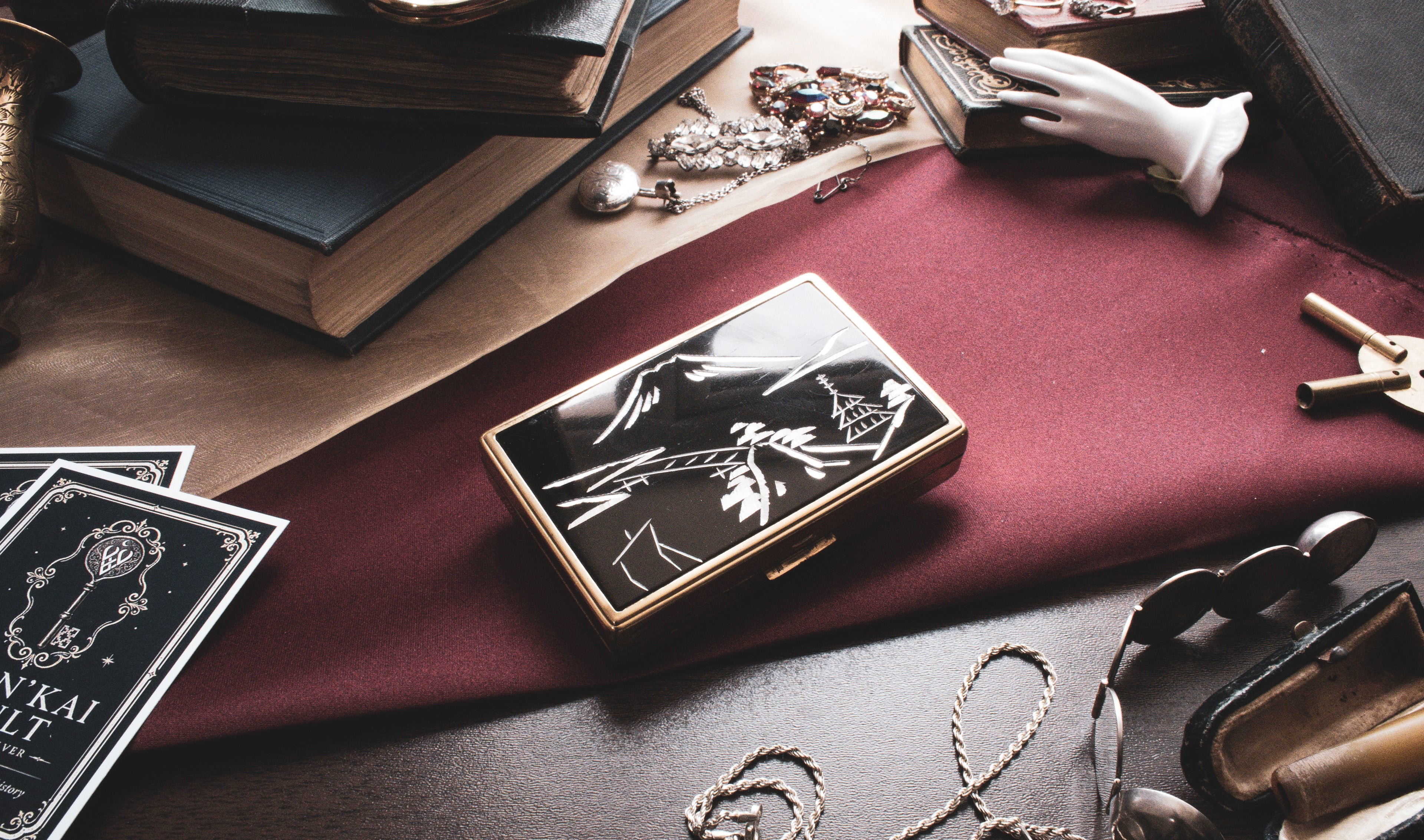 A rectangular black and gold case with an illustrated cover design, styled on a dark table alongside books, cards, jewelry, and other vintage props.