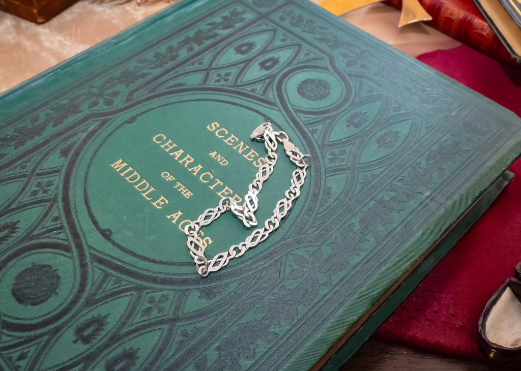 A silver-toned metal chain bracelet with a central heart-shaped link design, displayed on the cover of a vintage green book titled 'Scenes and Characters of the Middle Ages'.