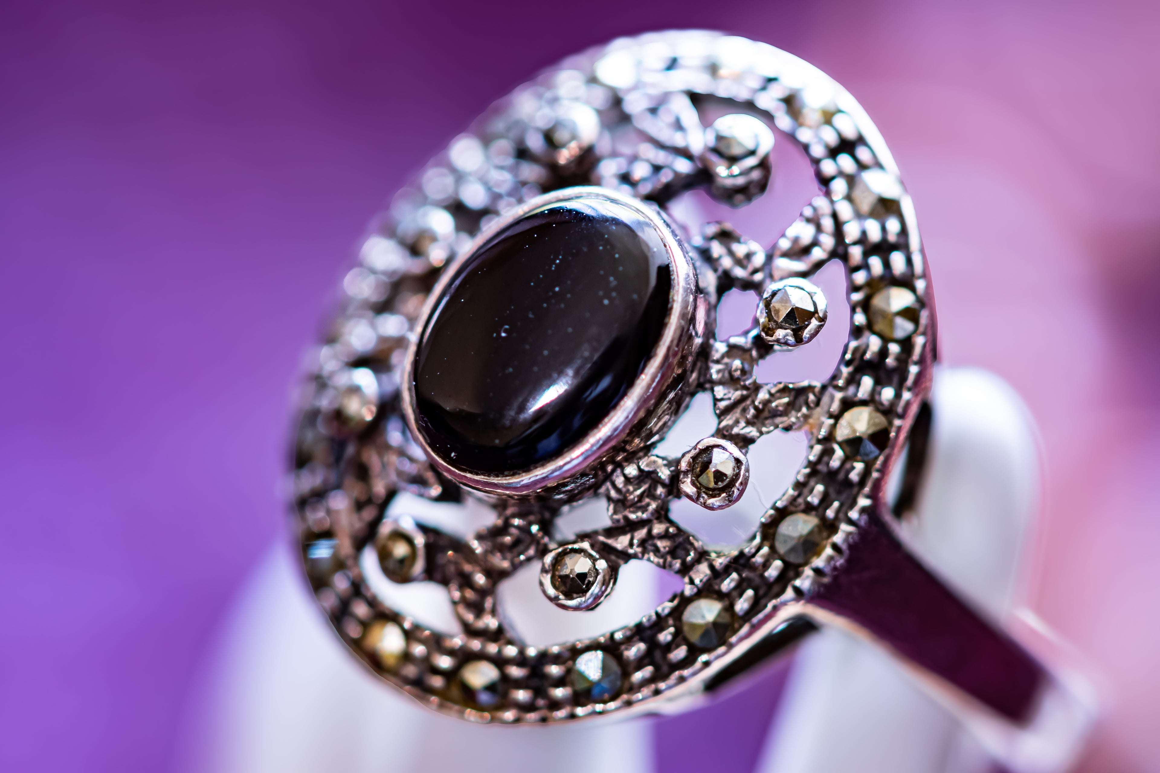 A close-up of an ornate silver ring with a large oval black cabochon stone and surrounding filigree detailing, held against a soft purple background.