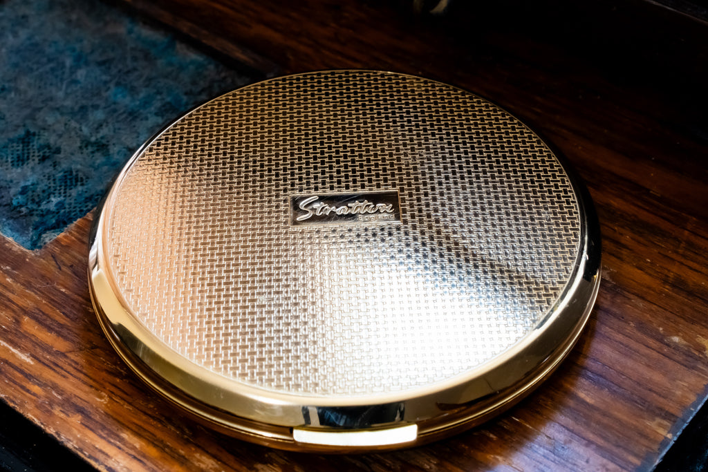 A round gold compact mirror with a textured honeycomb pattern lid and a small central 'Stratton' logo, resting on a dark wooden surface.