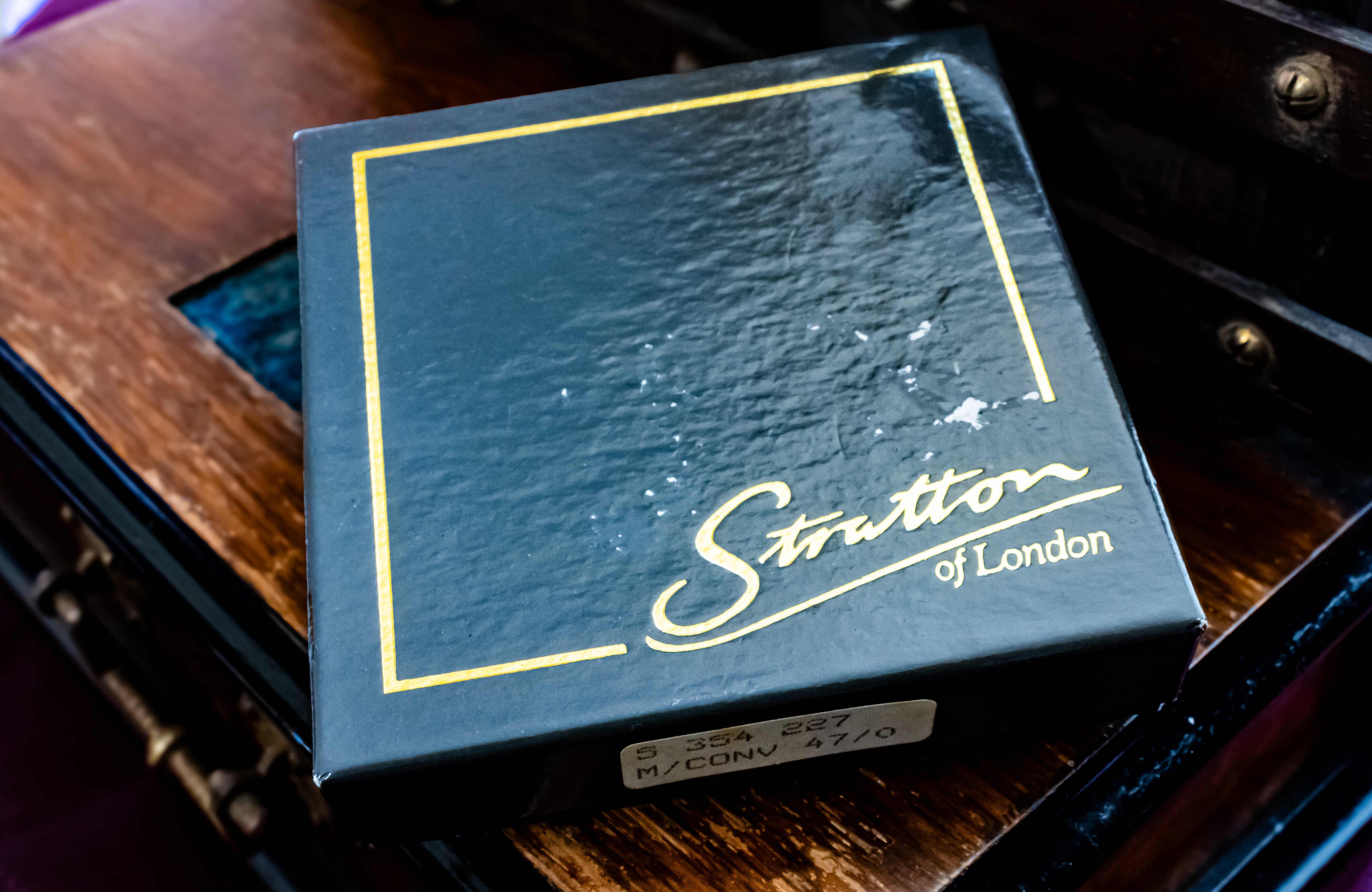 A close-up shot of a Stratton of London guitar string box, featuring a dark navy blue textured surface with gold foil borders and script lettering, resting on a dark wooden surface.