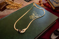 A gold chain necklace with a sculptural double-ring pendant, accented with black enamel and pavé crystals, displayed on a dark green leather-bound book.