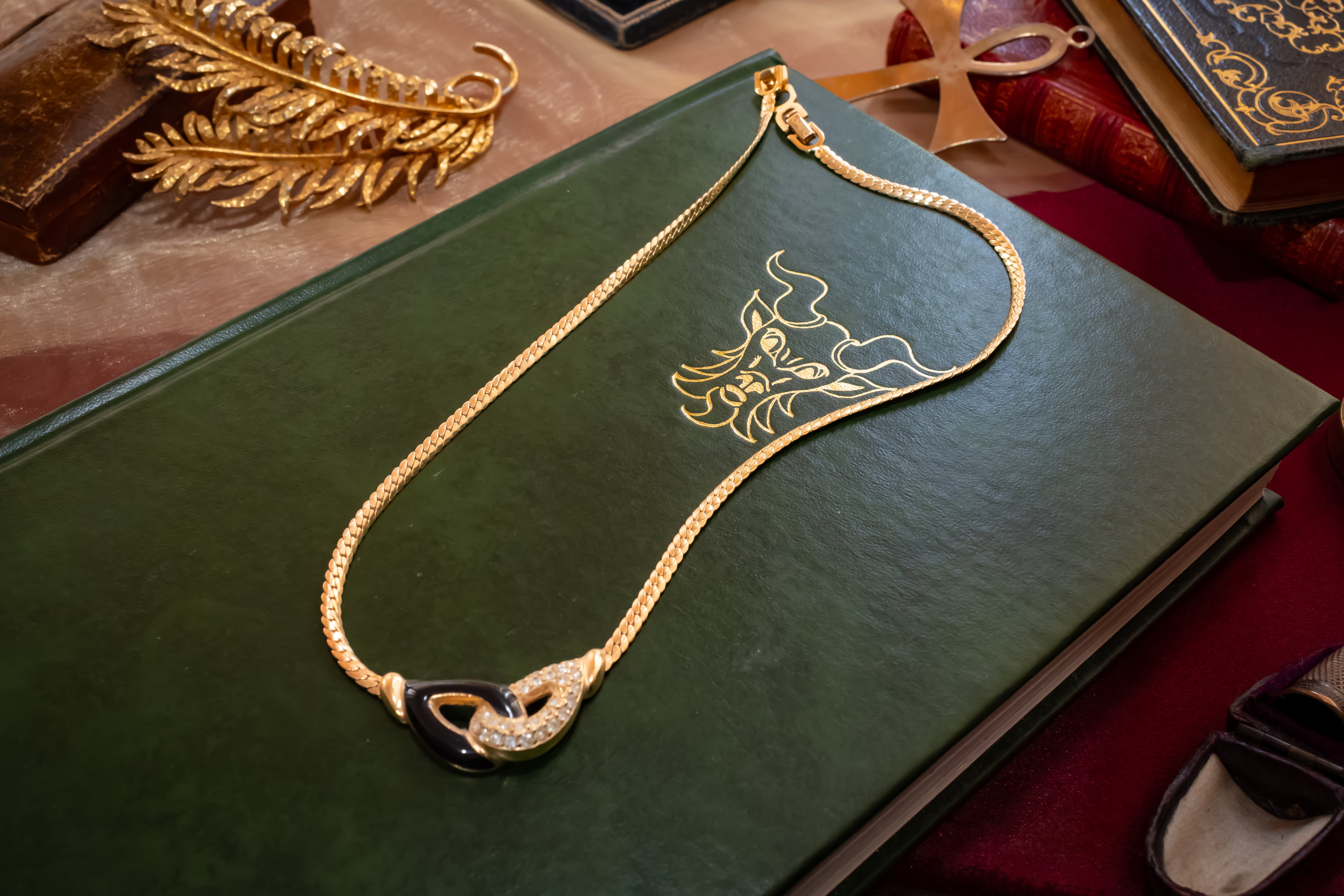 A gold chain necklace with a sculptural double-ring pendant, accented with black enamel and pavé crystals, displayed on a dark green leather-bound book.