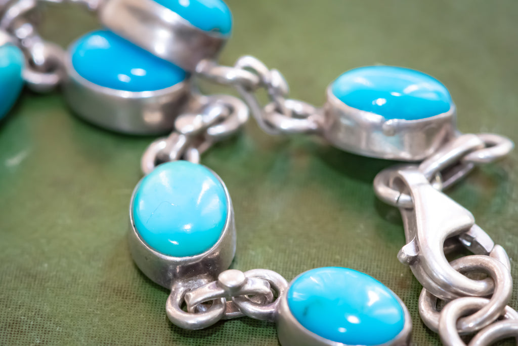 A close-up of a silver chain bracelet featuring oval turquoise cabochon stones set in silver bezels, resting on a textured green surface.