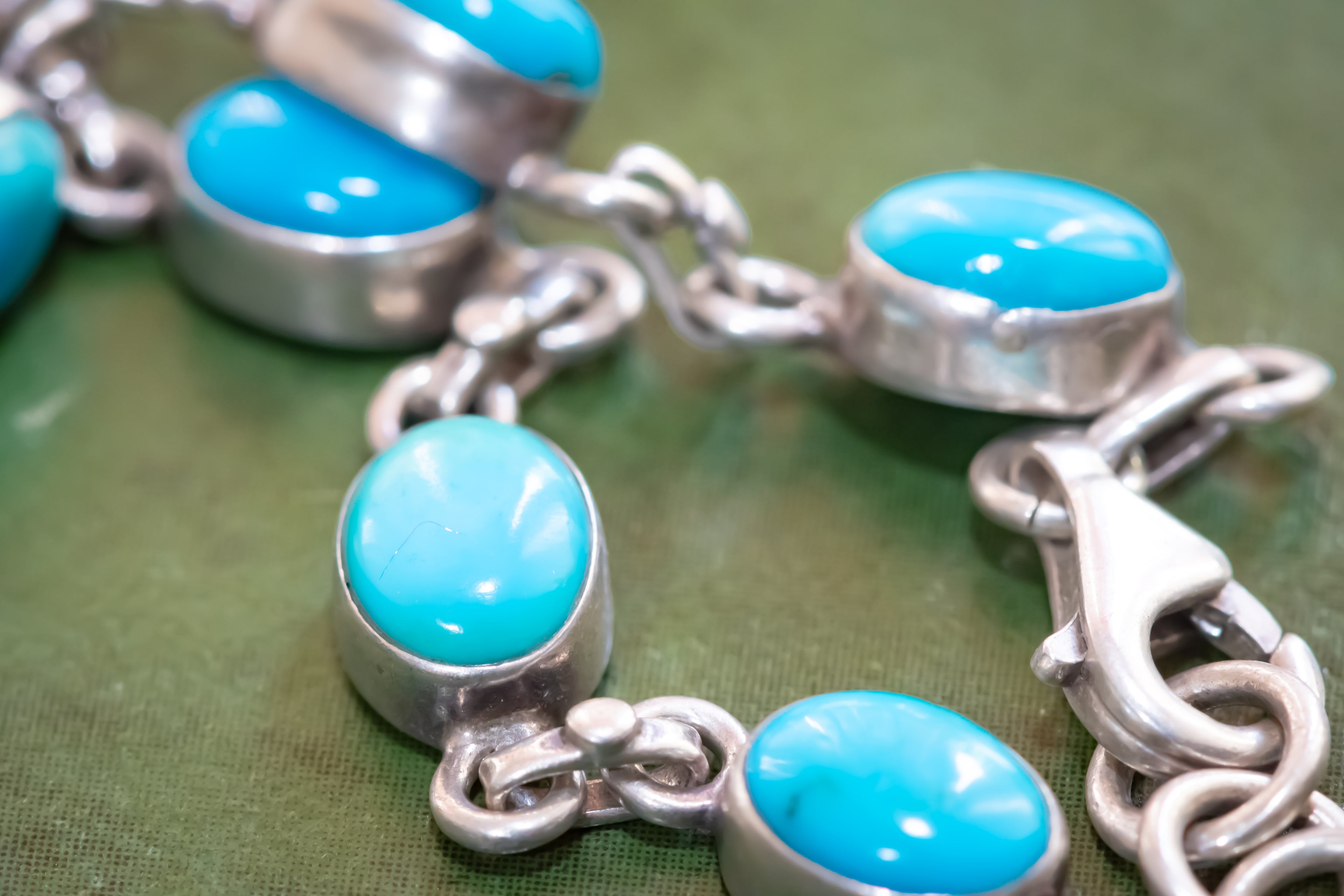 A close-up of a silver chain bracelet featuring oval turquoise cabochon stones set in silver bezels, resting on a textured green surface.