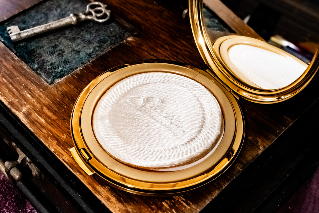 An open gold compact powder case with a white pressed powder and mirror, placed on a dark wooden surface next to a vintage key.