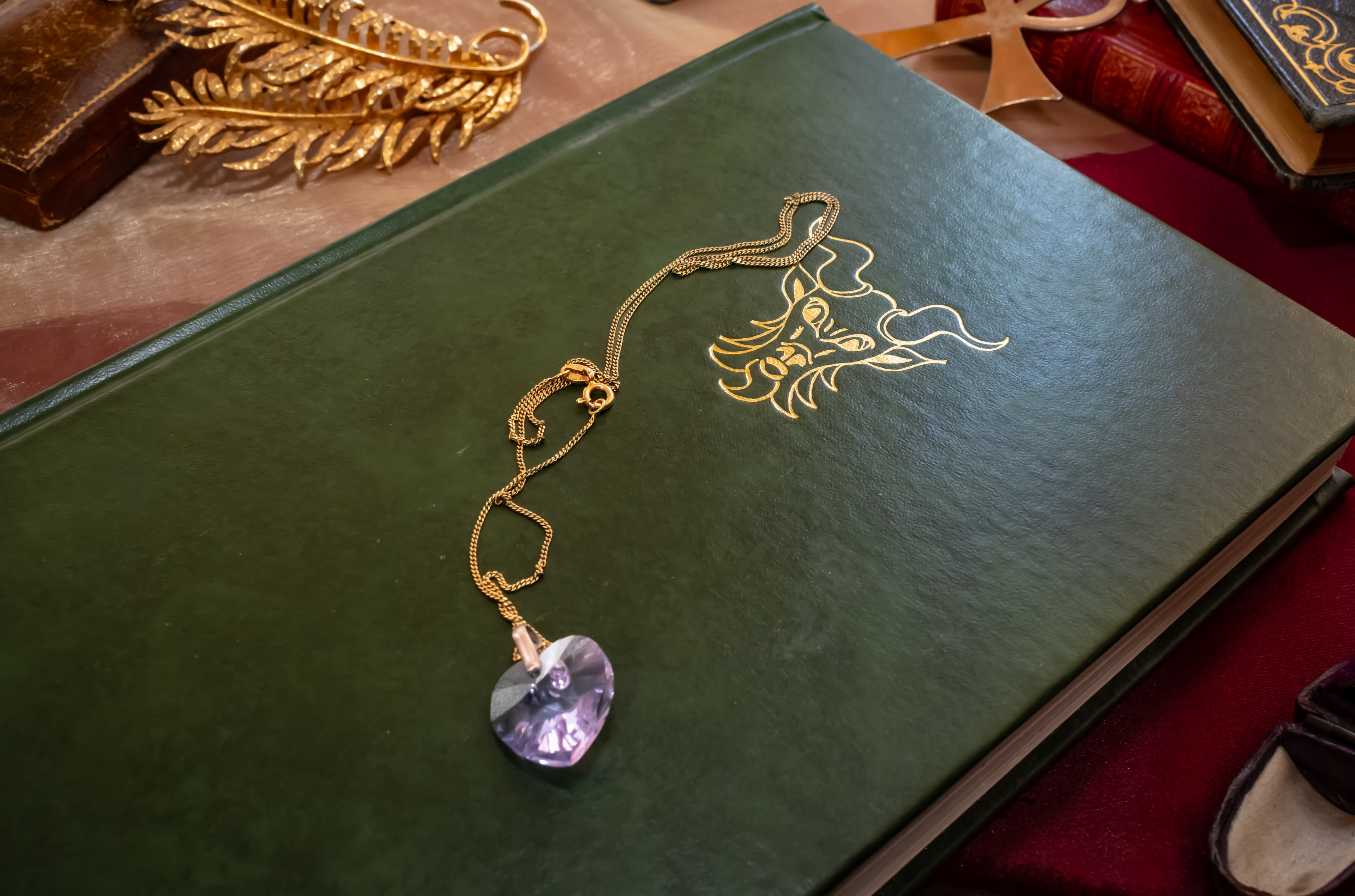 A gold-toned pendant necklace with a faceted purple heart-shaped gemstone, laid on a dark green book with gold foil decoration.