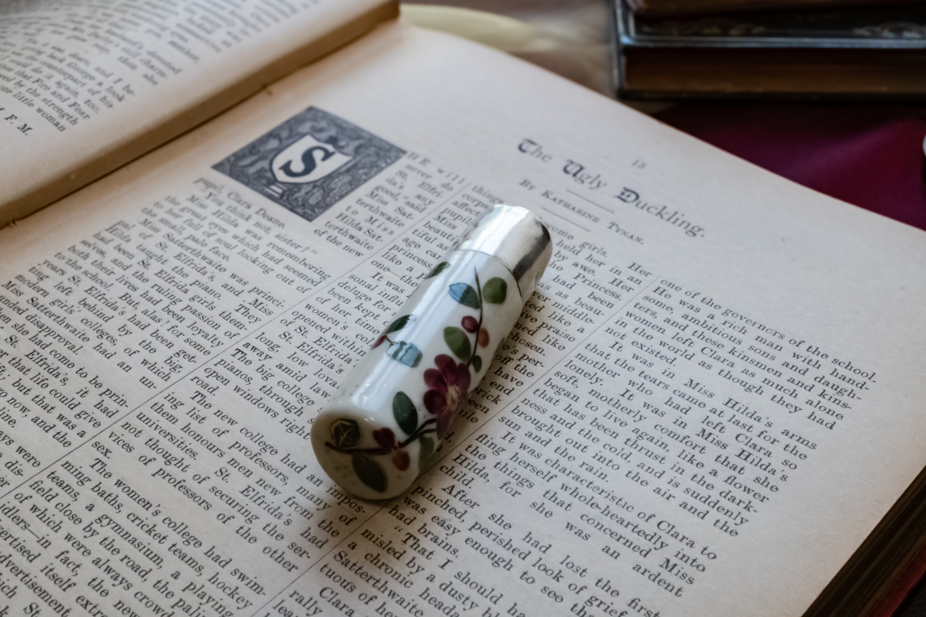 A vintage-style ceramic perfume bottle with a white body and a floral pattern of green leaves and red berries, resting on an open book page.