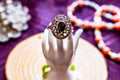 A silver-toned filigree ring with a large oval black stone, displayed on a white hand-shaped stand against a purple background with blurred jewelry props.