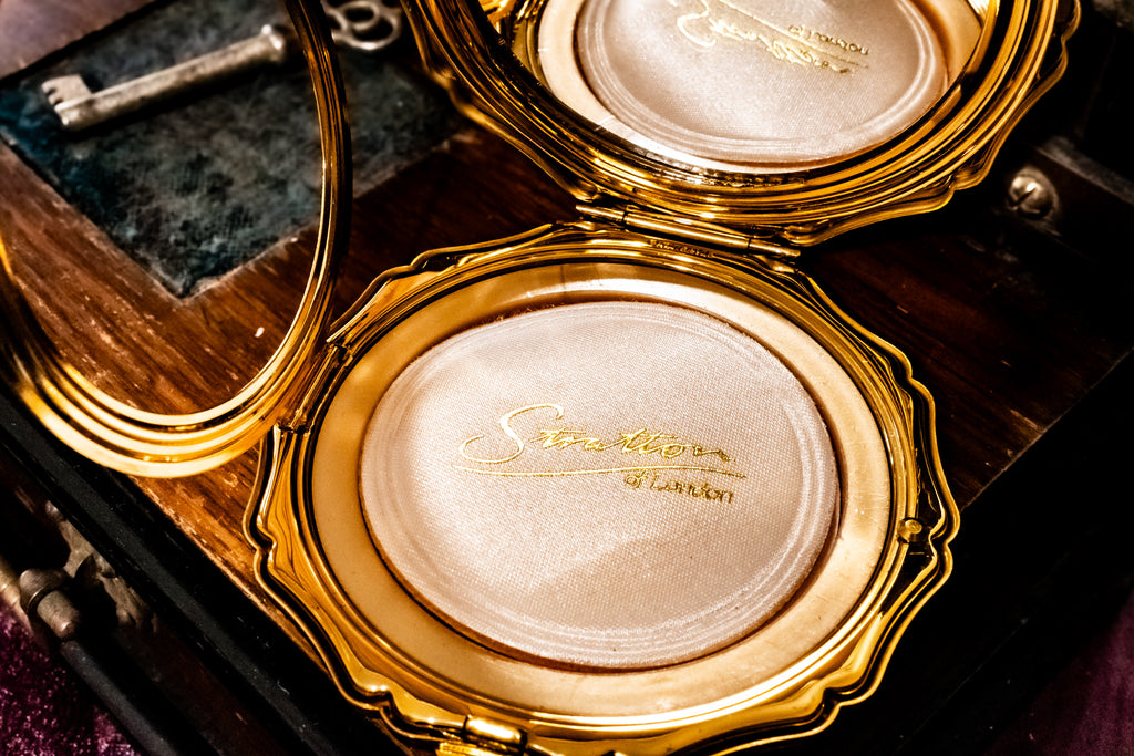 An ornate gold compact case opened on a dark wooden surface, showing a shimmering pale pink pressed powder with gold script reading 'Stratton of London' and a small mirror inside.