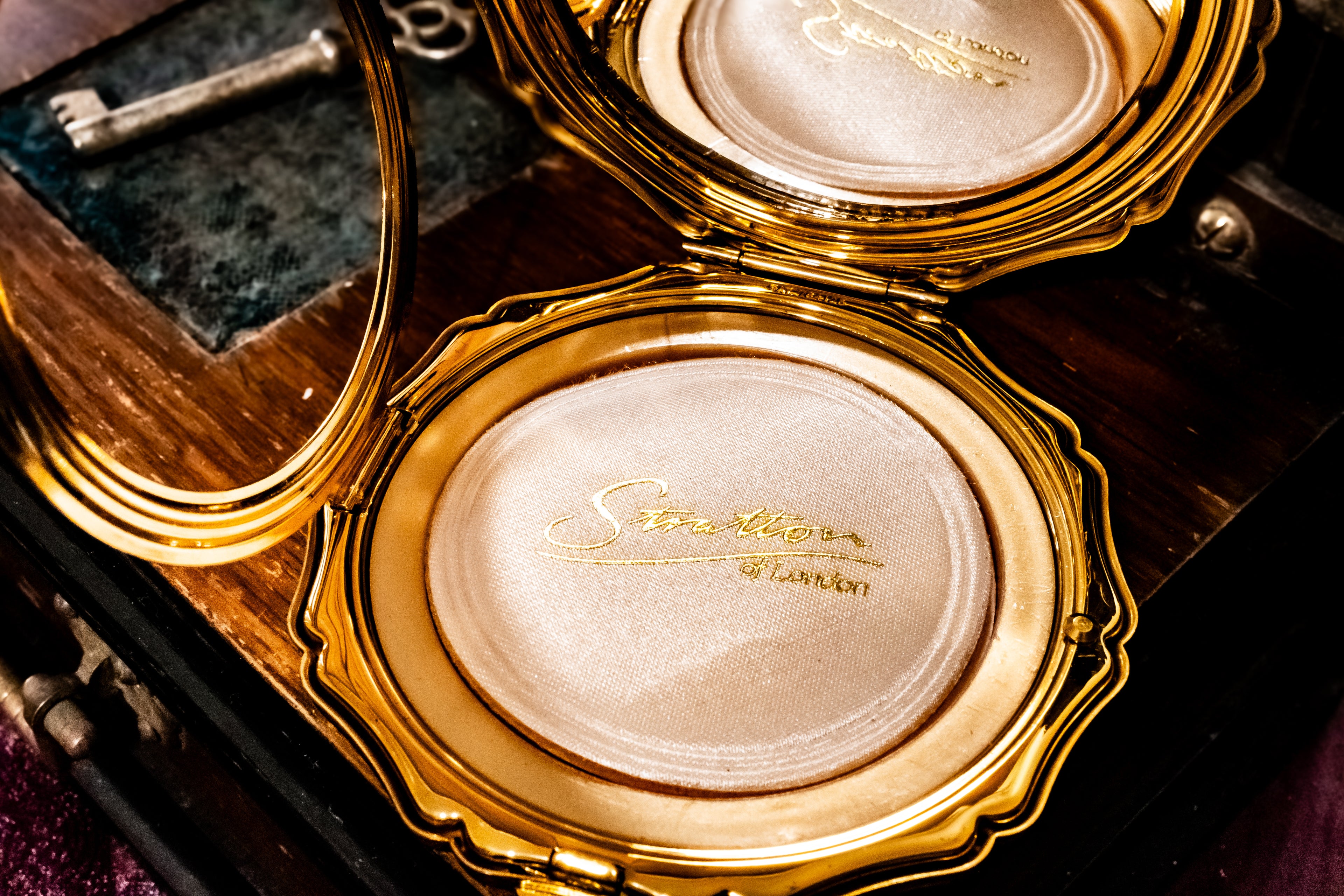 An ornate gold compact case opened on a dark wooden surface, showing a shimmering pale pink pressed powder with gold script reading 'Stratton of London' and a small mirror inside.