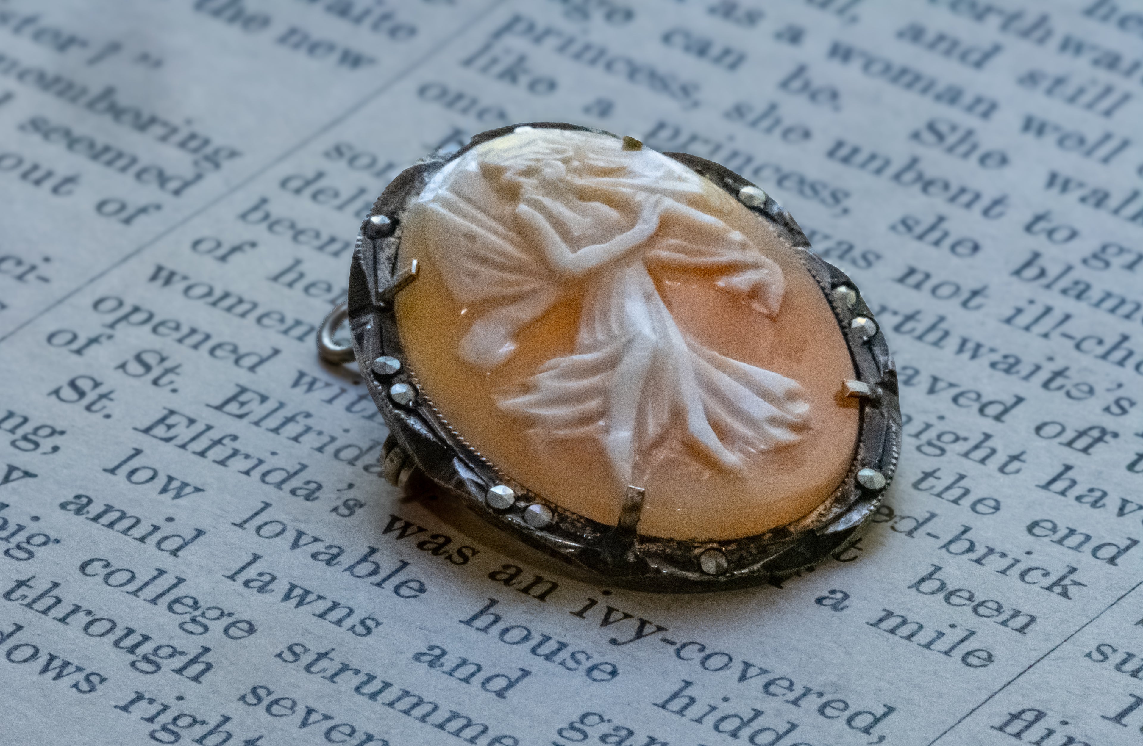 A vintage-style carved cameo pendant with a white figure design on a peach-colored oval background, set in an ornate silver-toned bezel with small clear stones, resting on printed book pages.