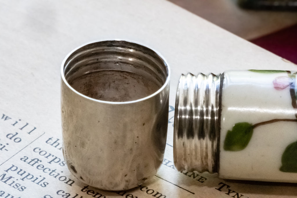 A close-up of a vintage ceramic perfume bottle with a silver metal lid, shown with the lid removed and placed beside it on printed paper.