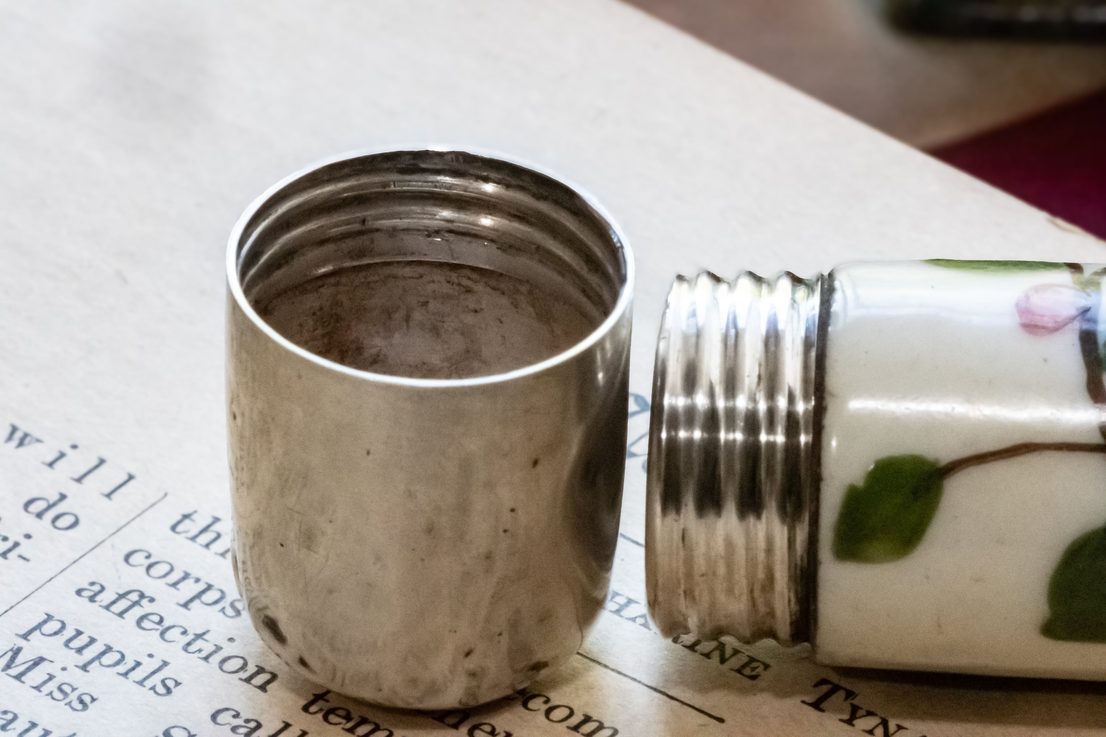 A close-up of a vintage ceramic perfume bottle with a silver metal lid, shown with the lid removed and placed beside it on printed paper.