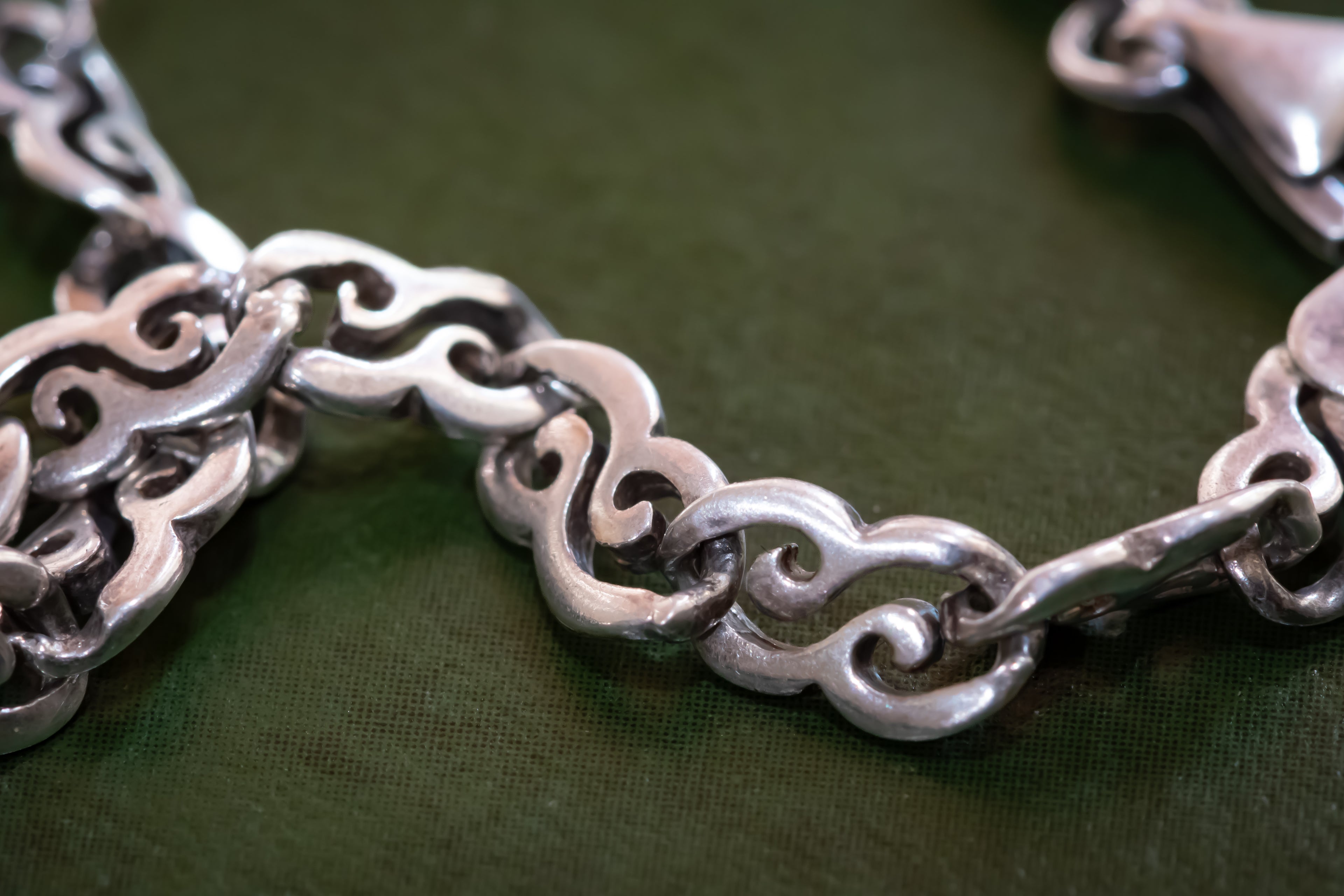 A close-up of a silver-toned chain bracelet with ornate interlocking links, resting on a dark olive green textured fabric background.