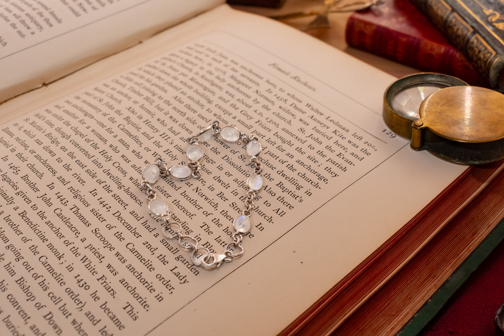 A silver-toned bracelet with round white stones is displayed on the pages of an open vintage book, surrounded by old books and a brass pocket watch.