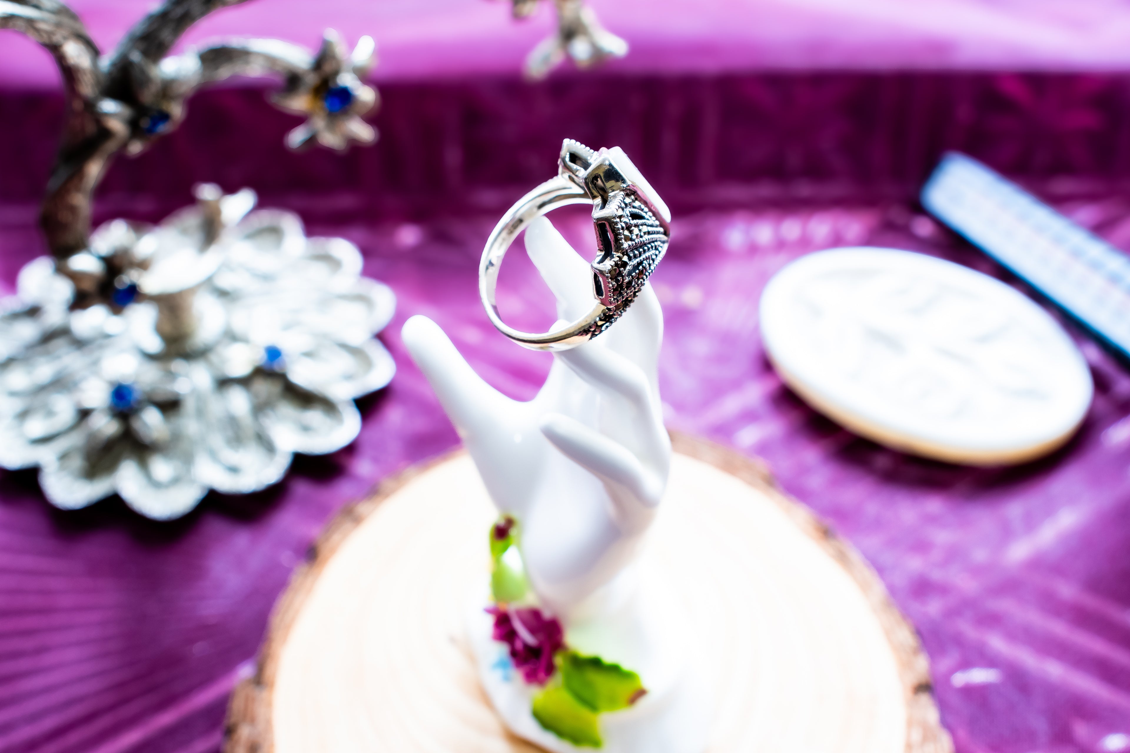 A silver ring with a detailed floral or wing-like setting, displayed on a white hand-shaped ring holder. The holder sits on a round wood slice, with a purple textured background and other decorative jewelry pieces blurred around it.
