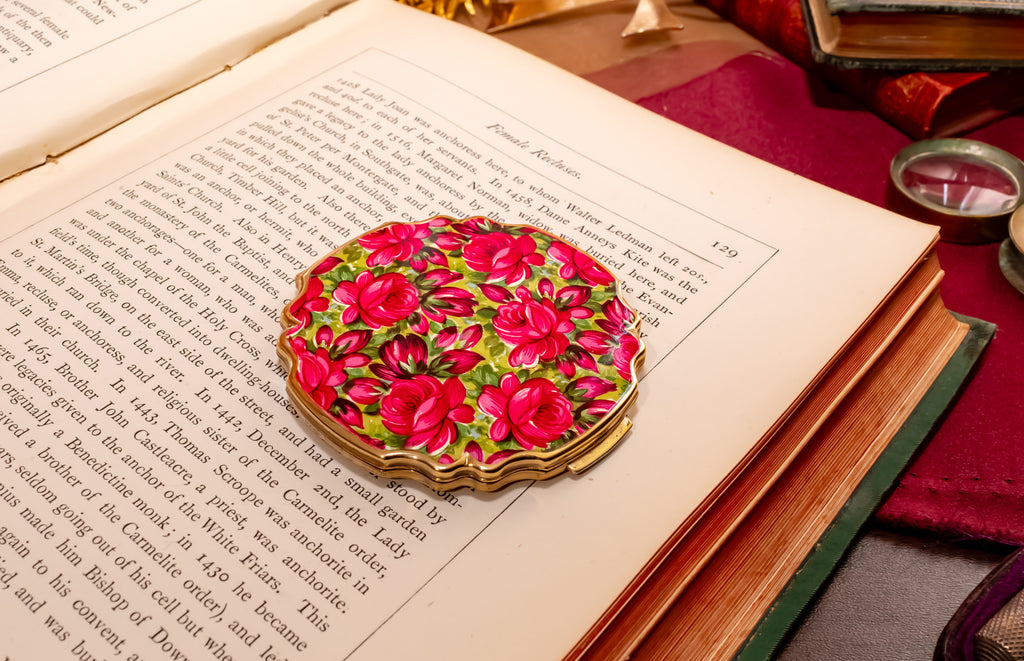 A round enamel Stratton powder compact with a vibrant pink rose floral pattern and gold scalloped edges, resting on an open vintage book.