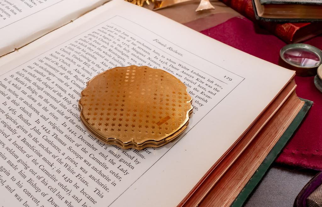 A round, gold-toned metal Stratton powder compact with a star etched bottom cover rests on the pages of an open vintage book, surrounded by old books and a red cloth.