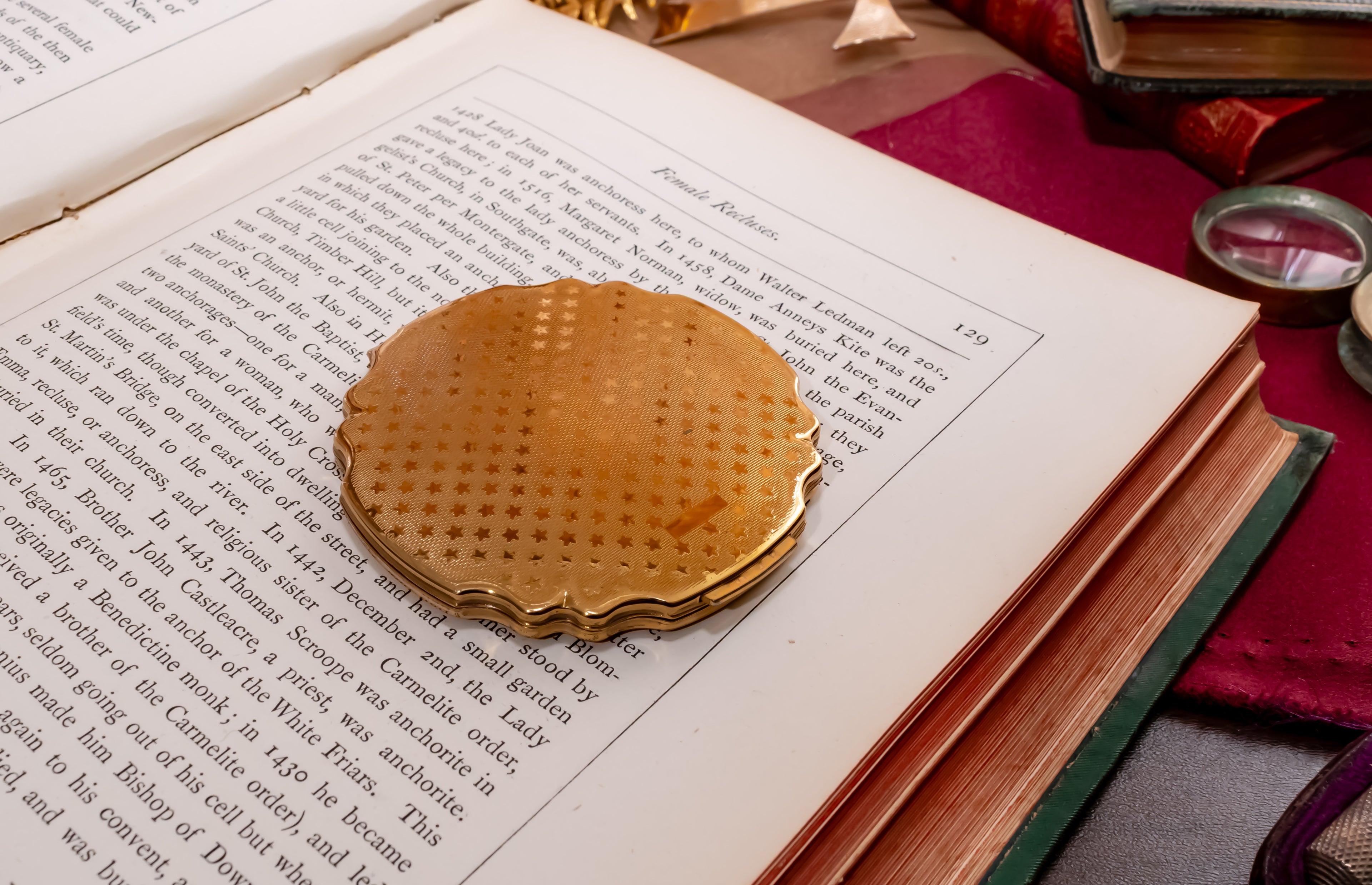 A round, gold-toned metal Stratton powder compact with a star etched bottom cover rests on the pages of an open vintage book, surrounded by old books and a red cloth.
