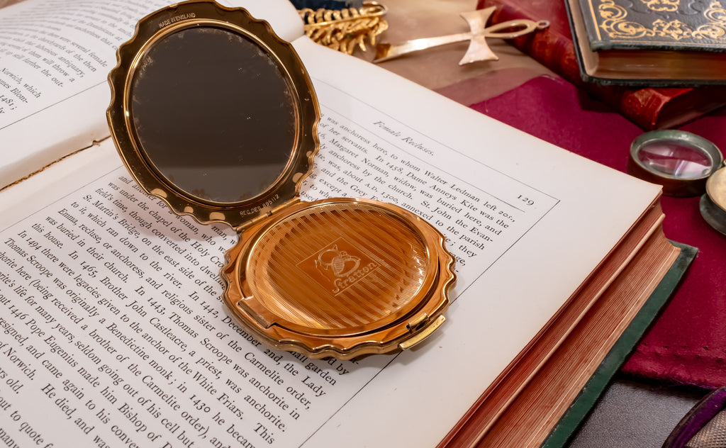A vintage gold tone Stratton powder compact, one open to show a dark reflective surface and one closed with a gold-leaf patterned interior, resting on the pages of an open book.