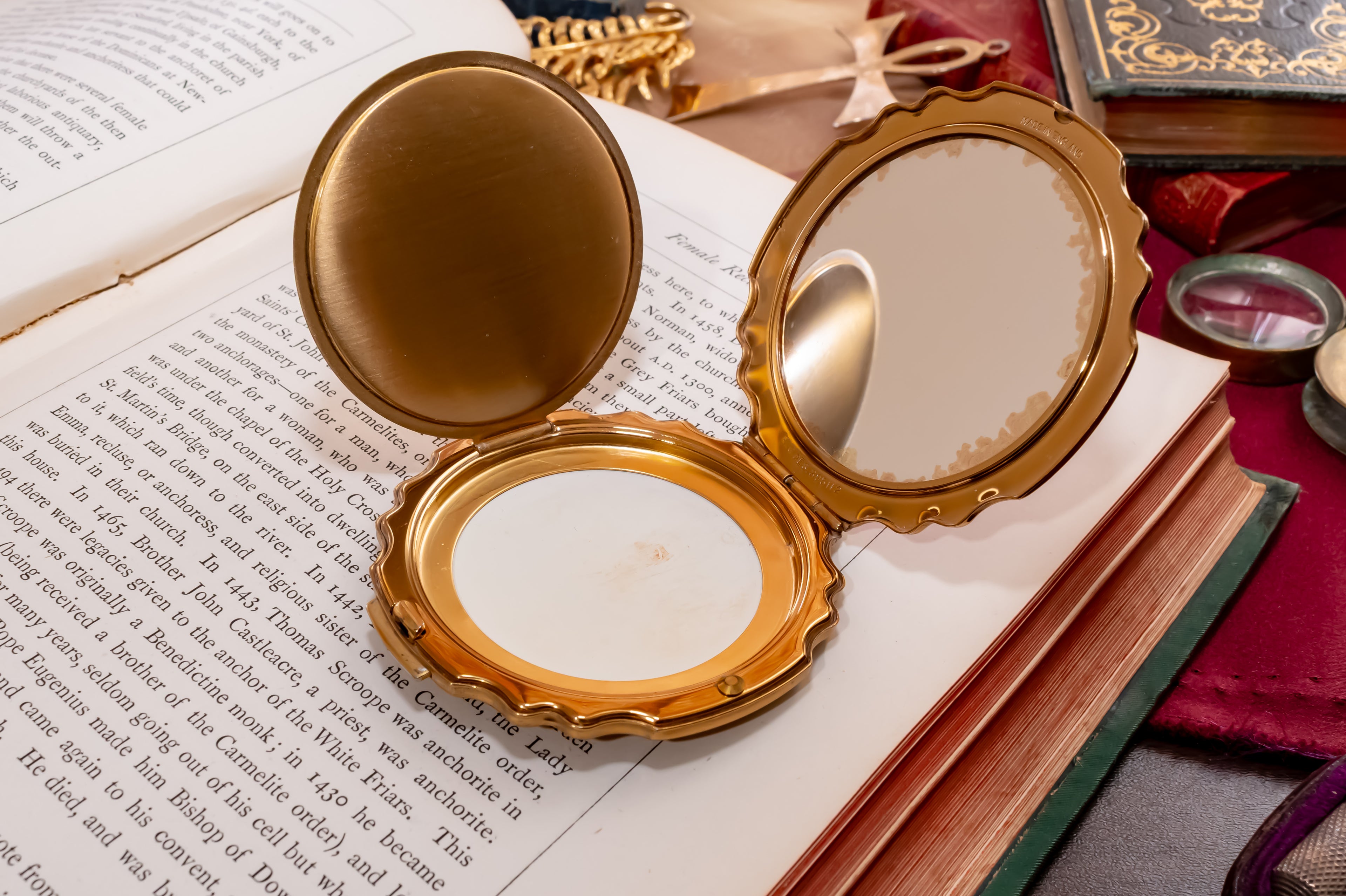 An ornate gold compact mirror with an open lid and a small mirror inside, placed on top of an open book surrounded by vintage books and a red cloth.