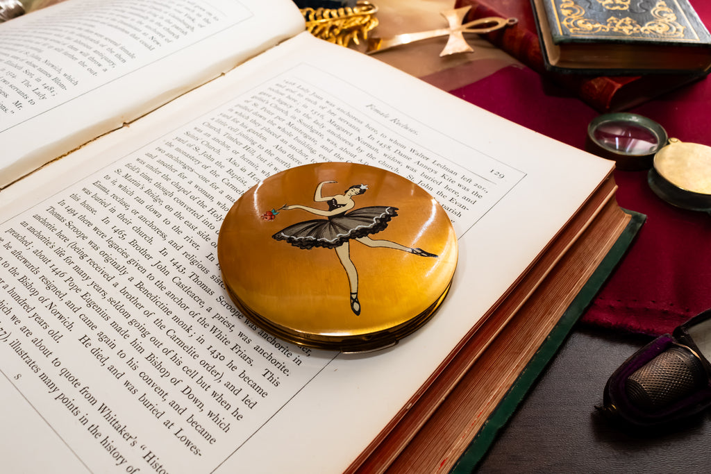 A round gold-toned Stratton compact mirror featuring an illustration of a ballerina in a black tutu, resting on an open book surrounded by vintage-style props.